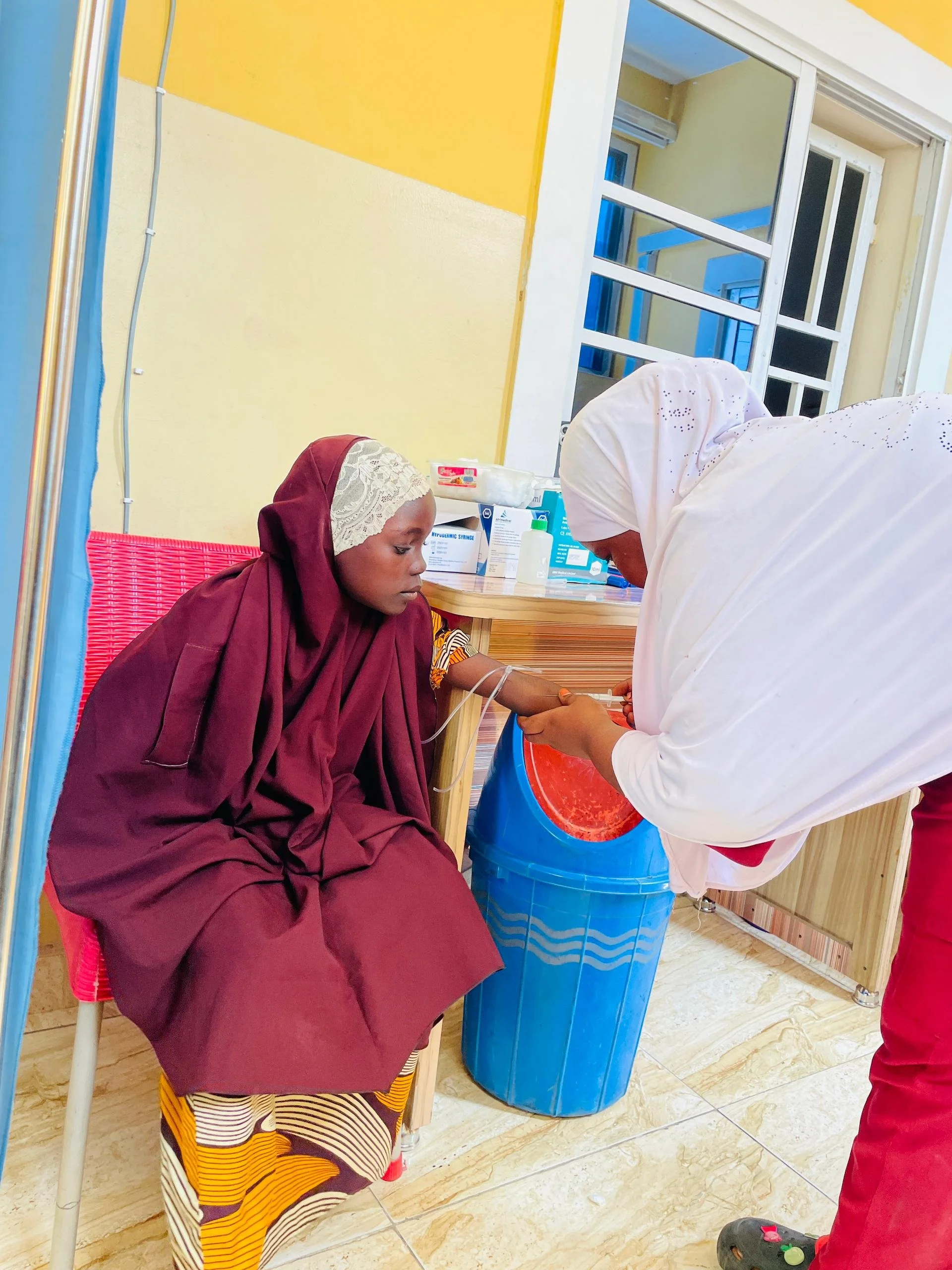 A woman in burgundy traditional clothing is sitting in a medical setting while a healthcare worker in white scrubs prepares to draw blood from her arm. The scene is inside a clinic with yellow walls and medical supplies on a counter.