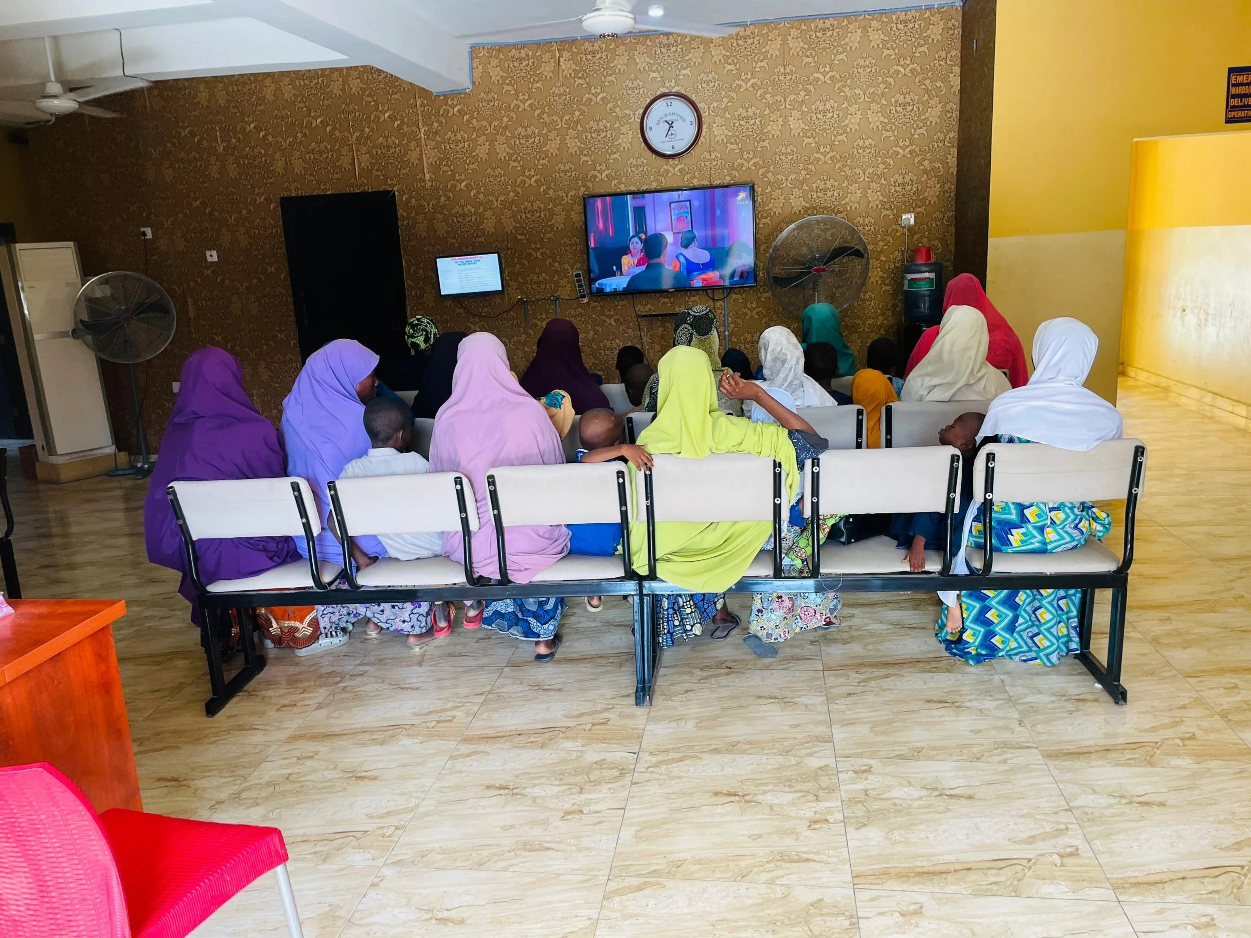 Group of women and children watching television in a room with gold and white patterned walls and wooden flooring.