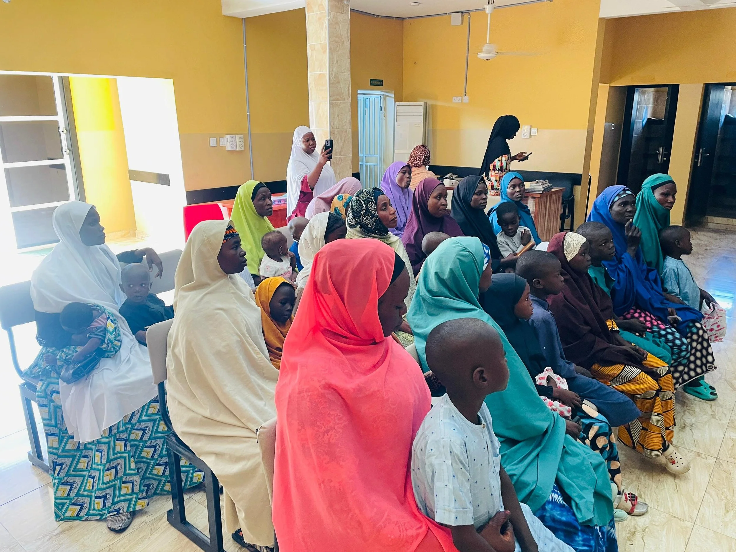 A group of women and children seated in a room, attending a gathering or event, with some women wearing colorful headscarves and modest clothing.