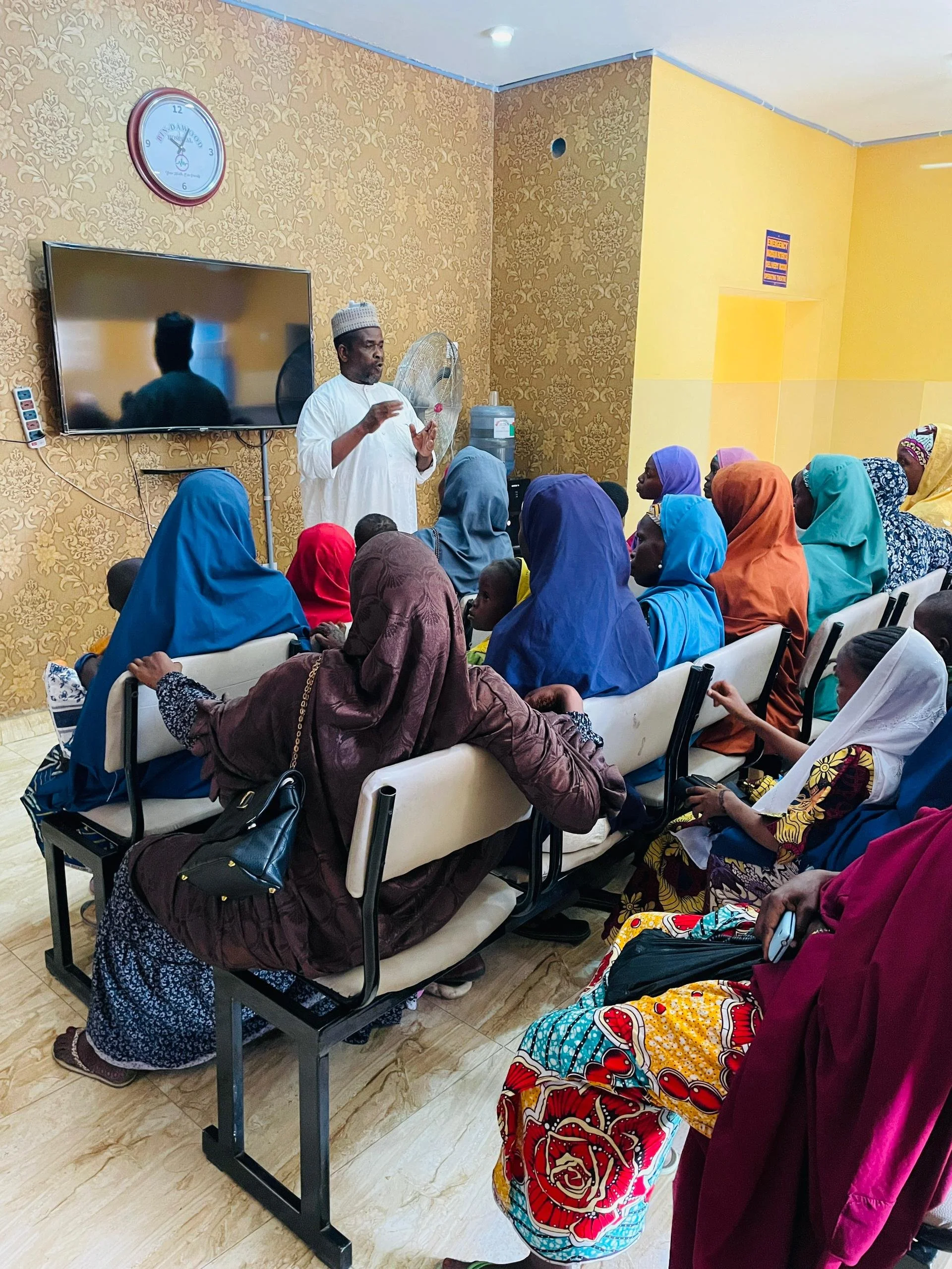 A man in white traditional attire and a white cap stands in front of a group of women and children in colorful clothing, giving a presentation or speech. The room has a decorative gold-patterned wallpaper, a wall clock, and a flat-screen TV on the wa
