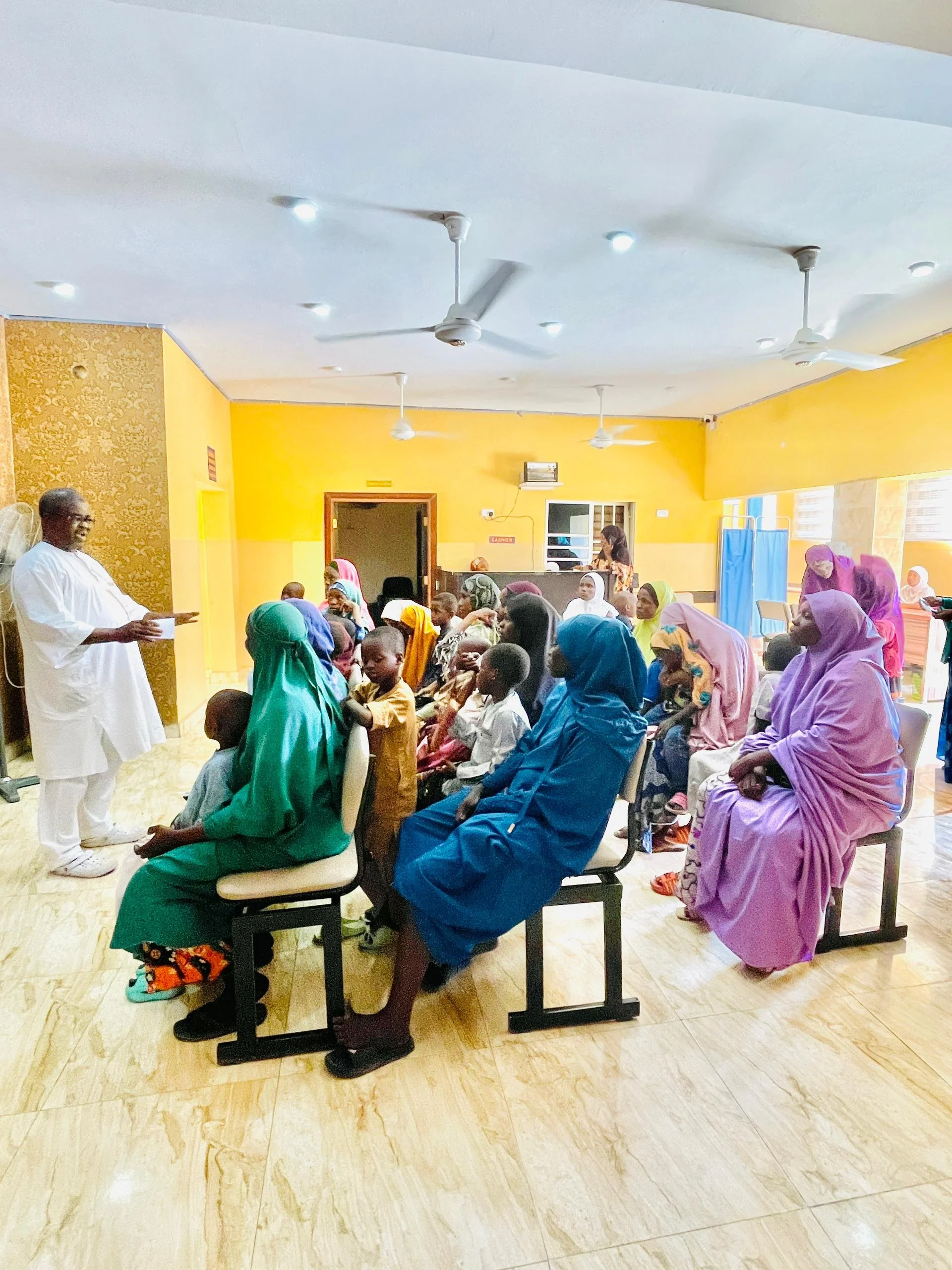 A group of people, including children and women wearing colorful traditional attire, seated in a hall listening to a man standing at the front, possibly giving a speech or presentation. The hall has a bright yellow wall and a wooden floor.