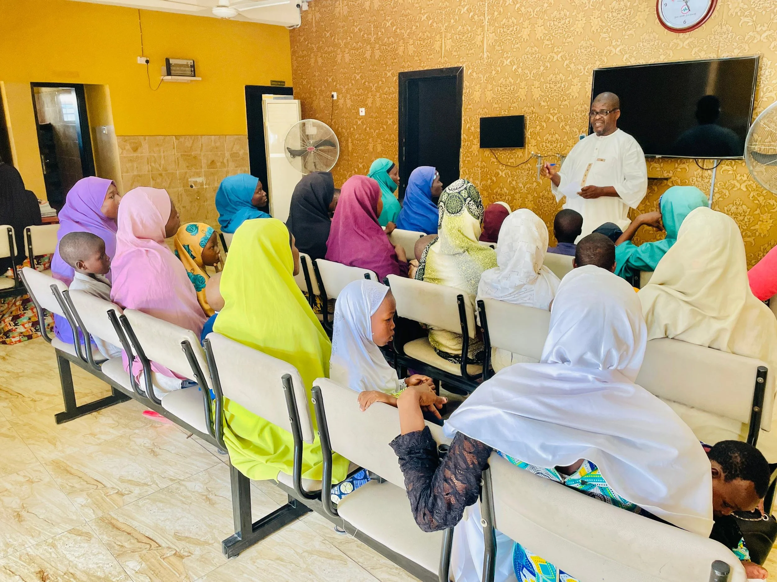A group of people, mostly women wearing colorful headscarves, sitting on chairs in a room, listening to a man in white speaking at a front. The room has yellow and gold decor, a large black television, and two fans.