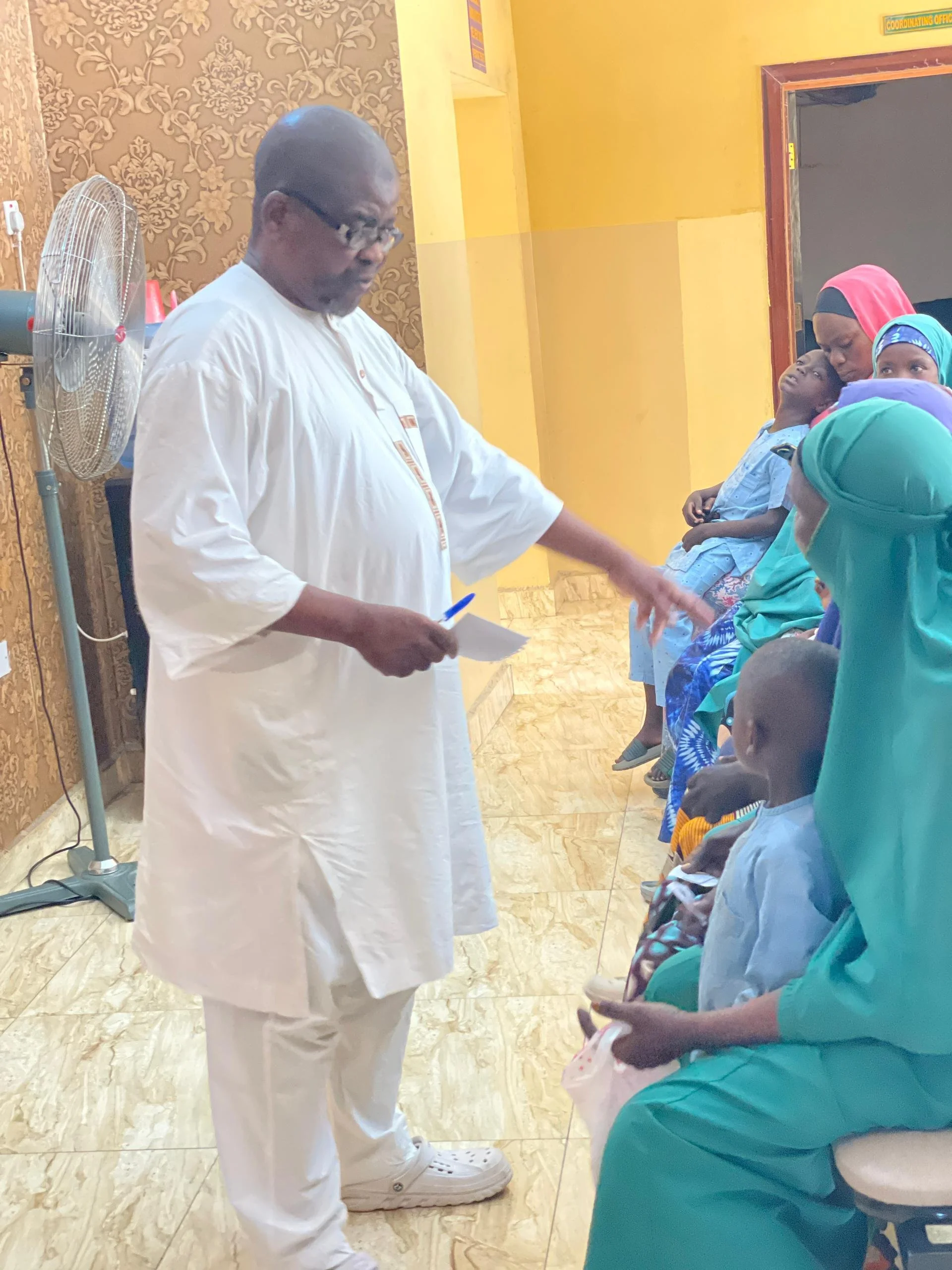 A man in white traditional attire standing in front of a group of children and women, giving a presentation or talk in a room with yellow walls and a patterned wall on the left, with some children looking up and listening attentively.