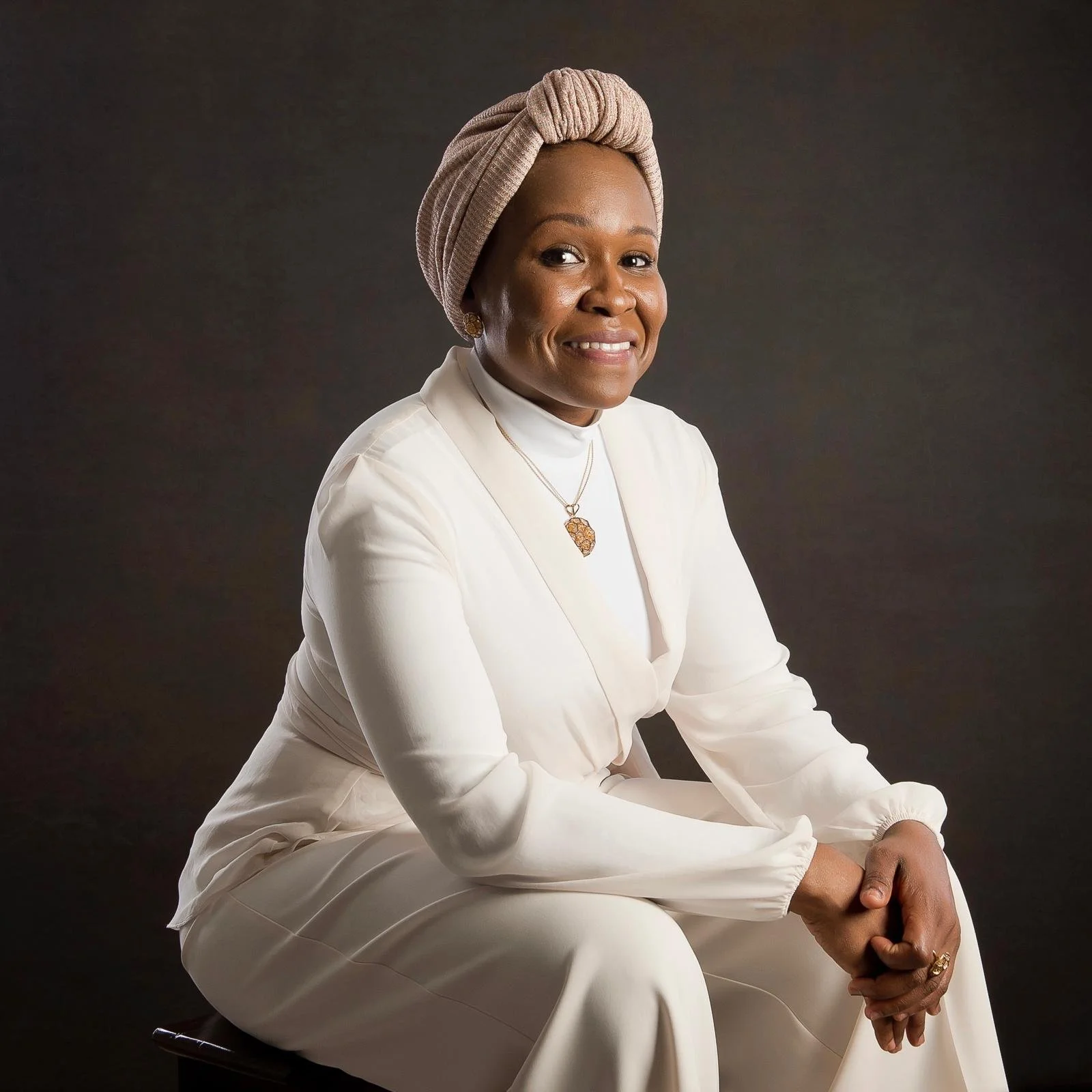 A woman in white attire sitting on a stool against a dark background, wearing a headwrap and jewelry, smiling at the camera.