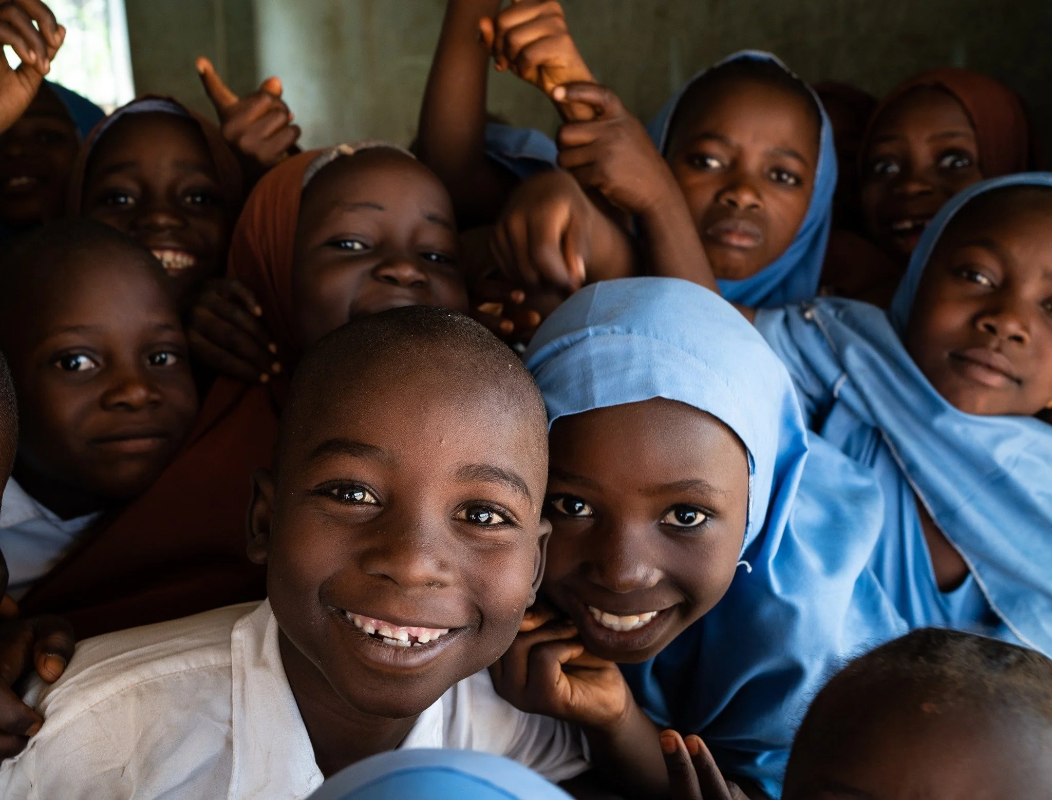 Group of smiling children, some in school uniforms, gathered closely together indoors.