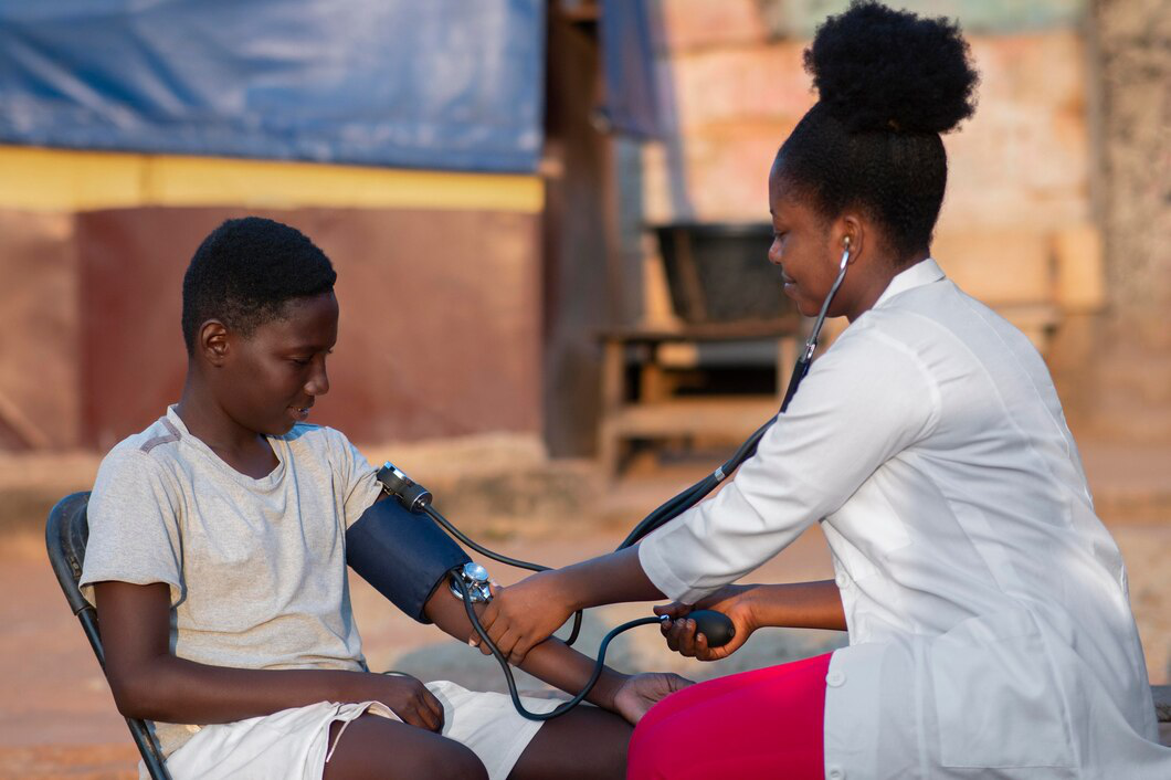 A healthcare worker in a white coat measures the blood pressure of a boy sitting on a chair outdoors using a sphygmomanometer.