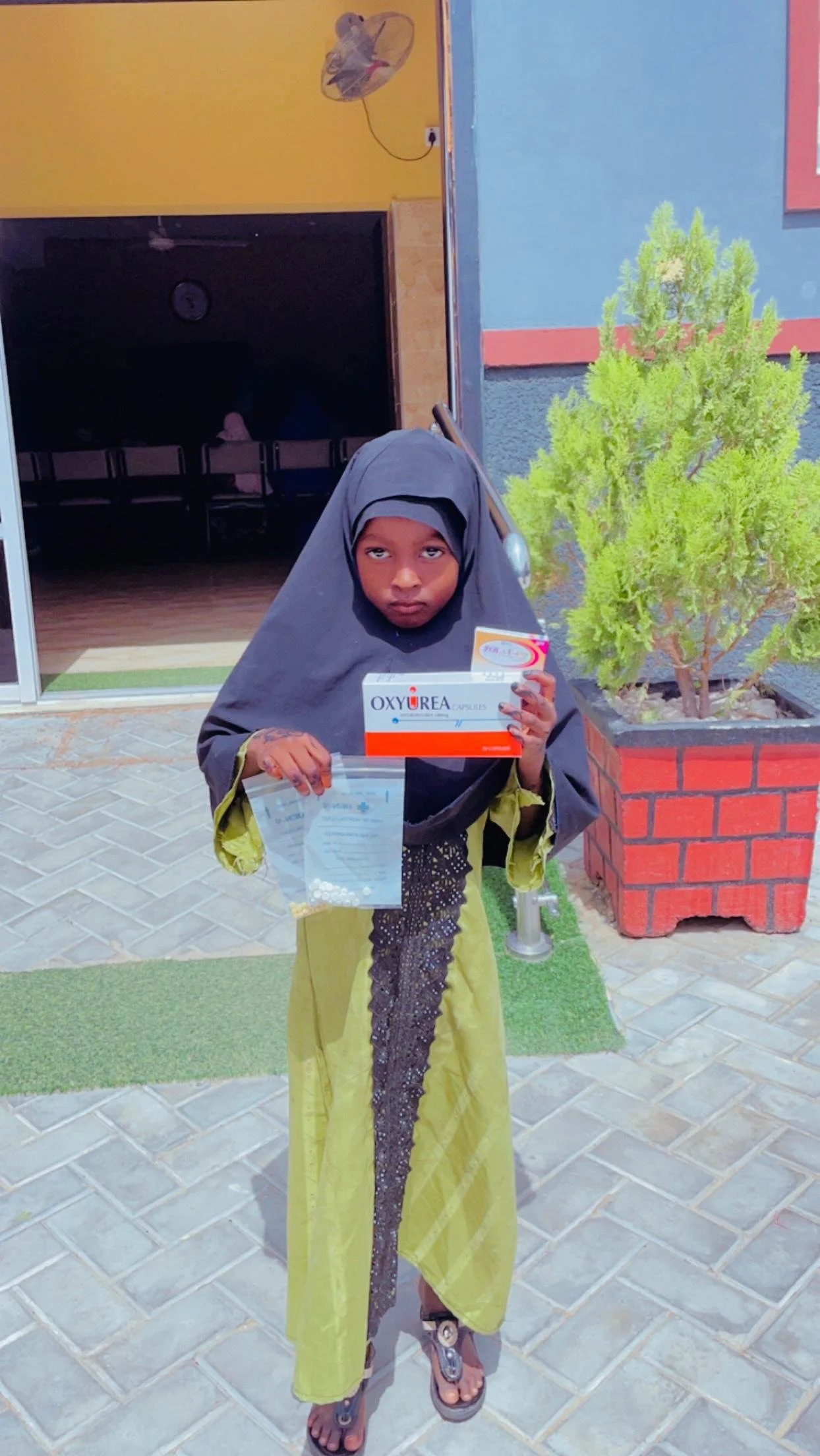 Young girl wearing a black headscarf and a green dress, standing outside near a potted plant, holding a box labeled 'Oxyurea' and a plastic bag.