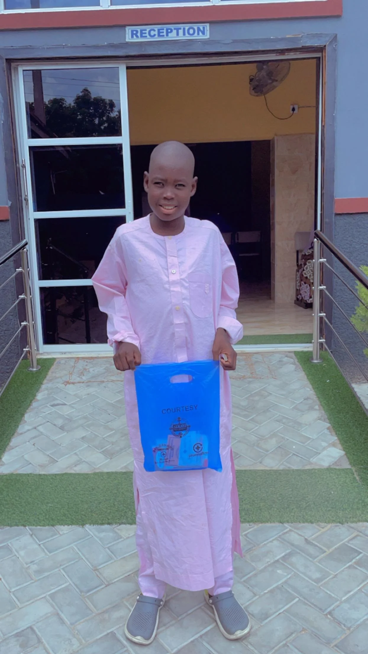 A young boy with a shaved head standing outside a building labeled 'Reception', holding a blue plastic bag, wearing a light pink traditional outfit and grey slip-on shoes.