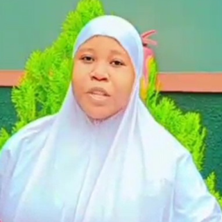 A woman wearing a white hijab sitting outdoors with green foliage in the background.