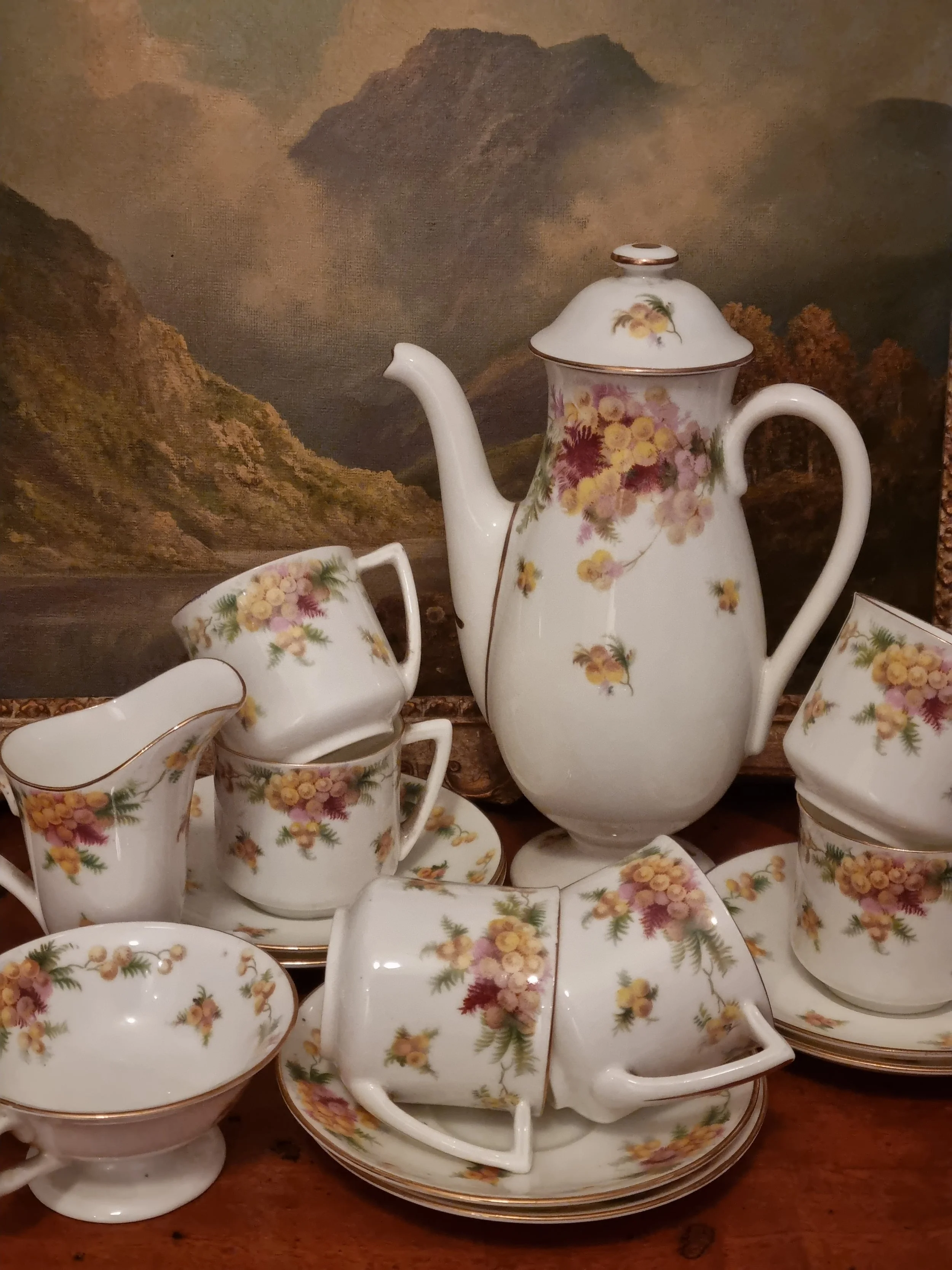 Set of floral patterned porcelain teacups, saucers, creamer, and teapot with a mountain landscape painting in the background.