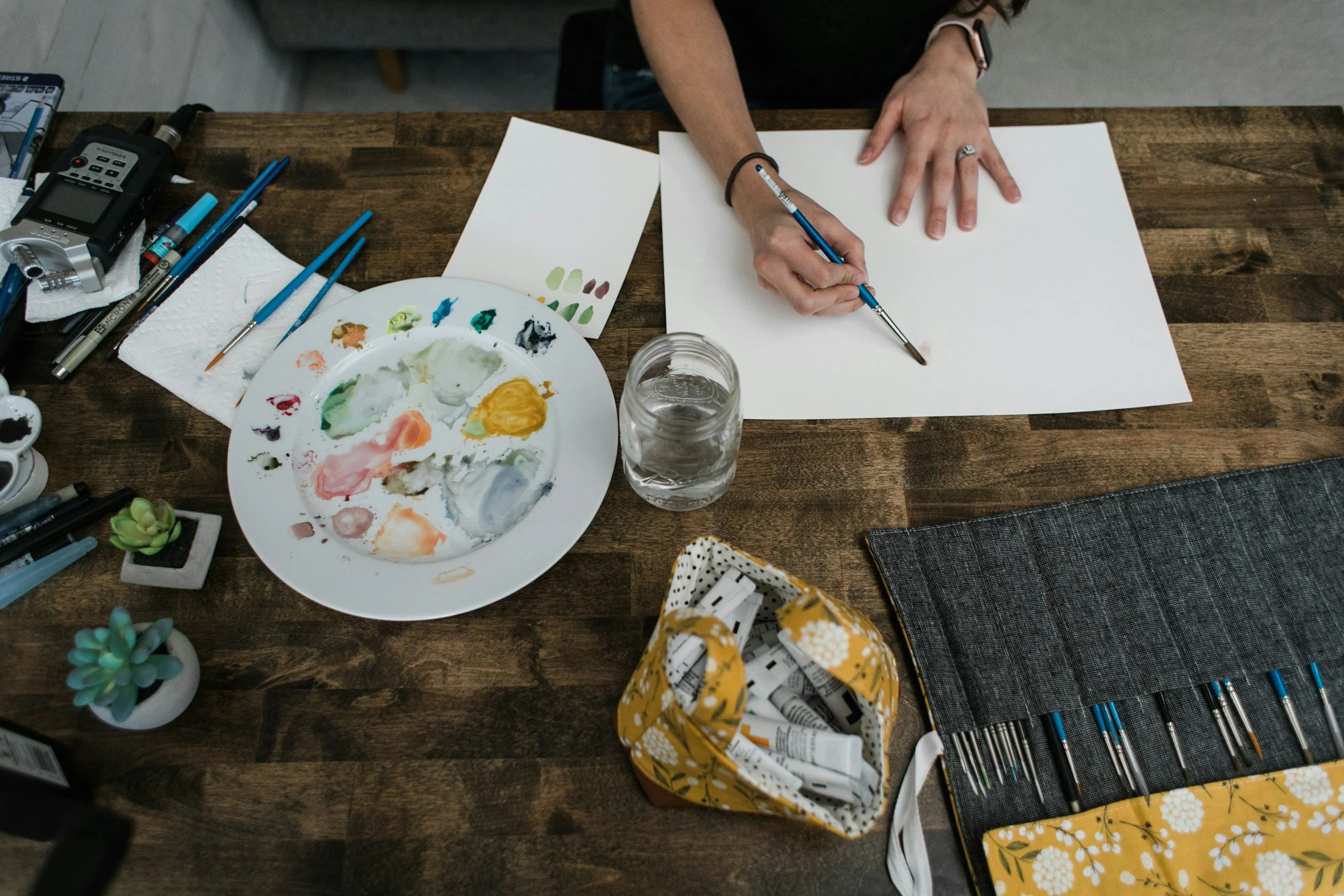 A person painting with watercolors on a blank white sheet of paper on a wooden table. The table has art supplies, including brushes, a palette with colored paints, a glass of water, and a potted succulent plant.