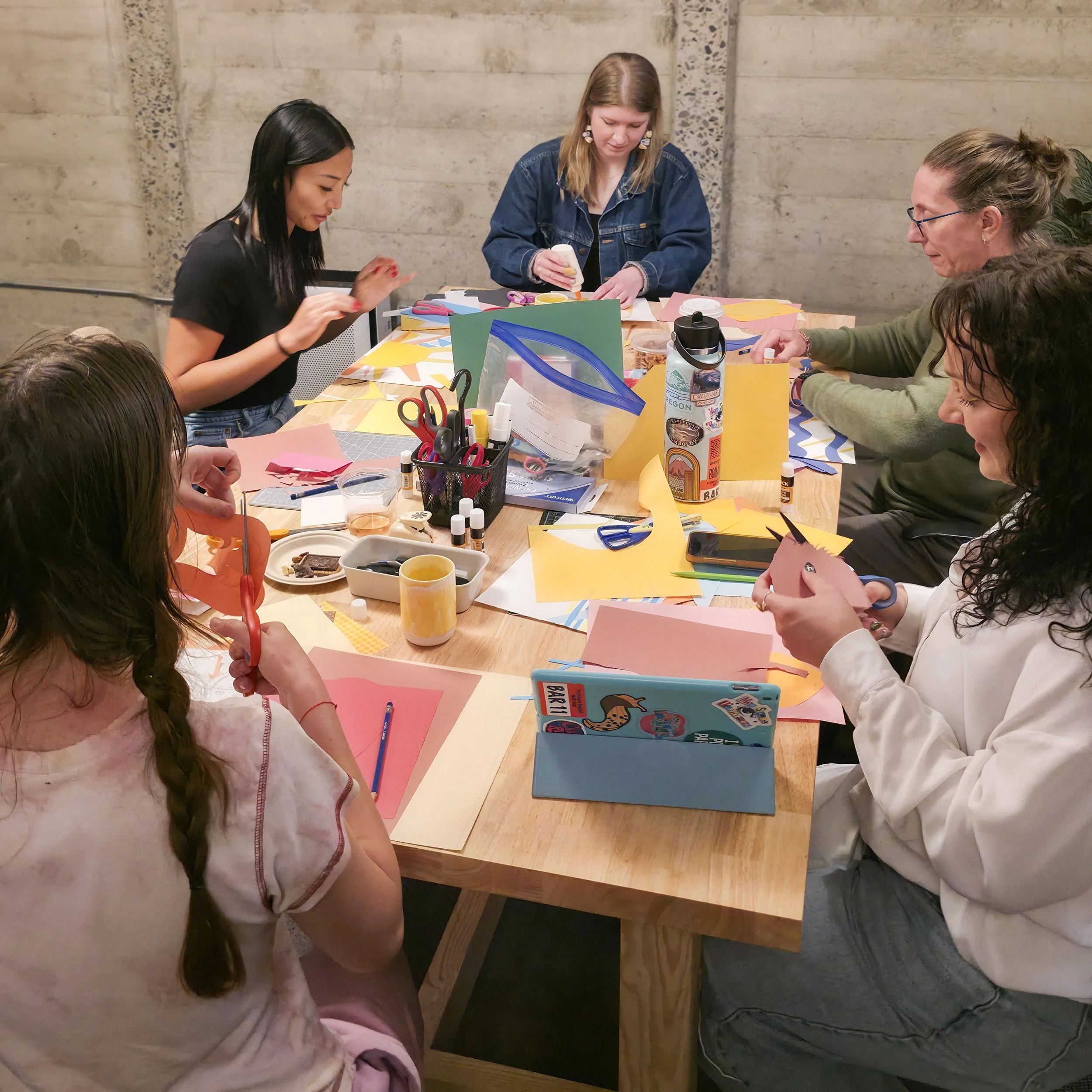 Group of people creating colorful paper crafts together at a workshop table with scissors, glue, markers, and art supplies.
