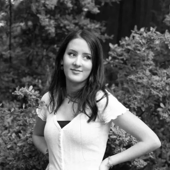 A young woman with shoulder-length dark hair, wearing a light-colored short-sleeve top and a necklace, standing outdoors in front of bushes and trees.