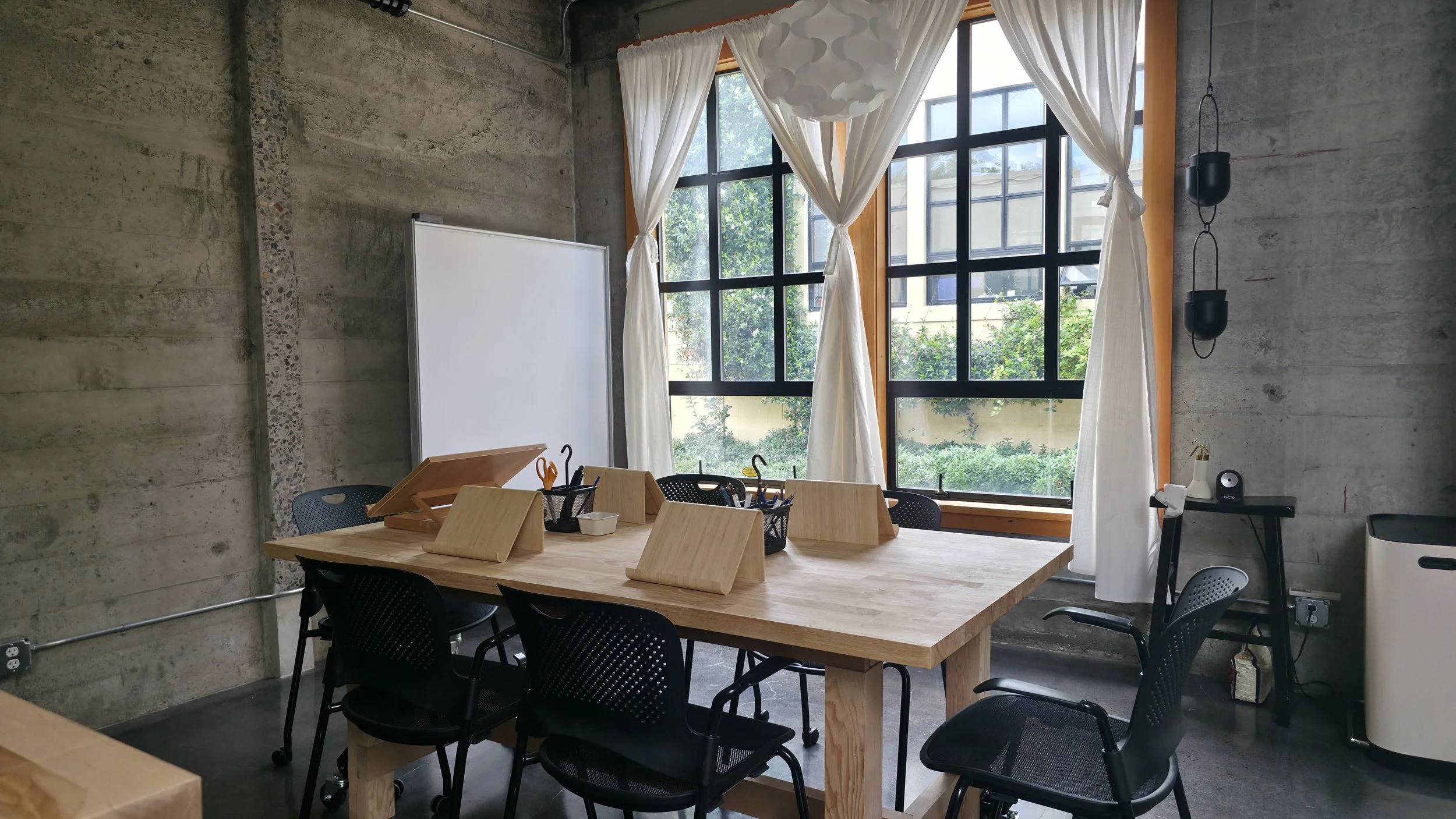 A modern office space with a wooden table, black chairs, large window with white curtains, and a concrete wall.