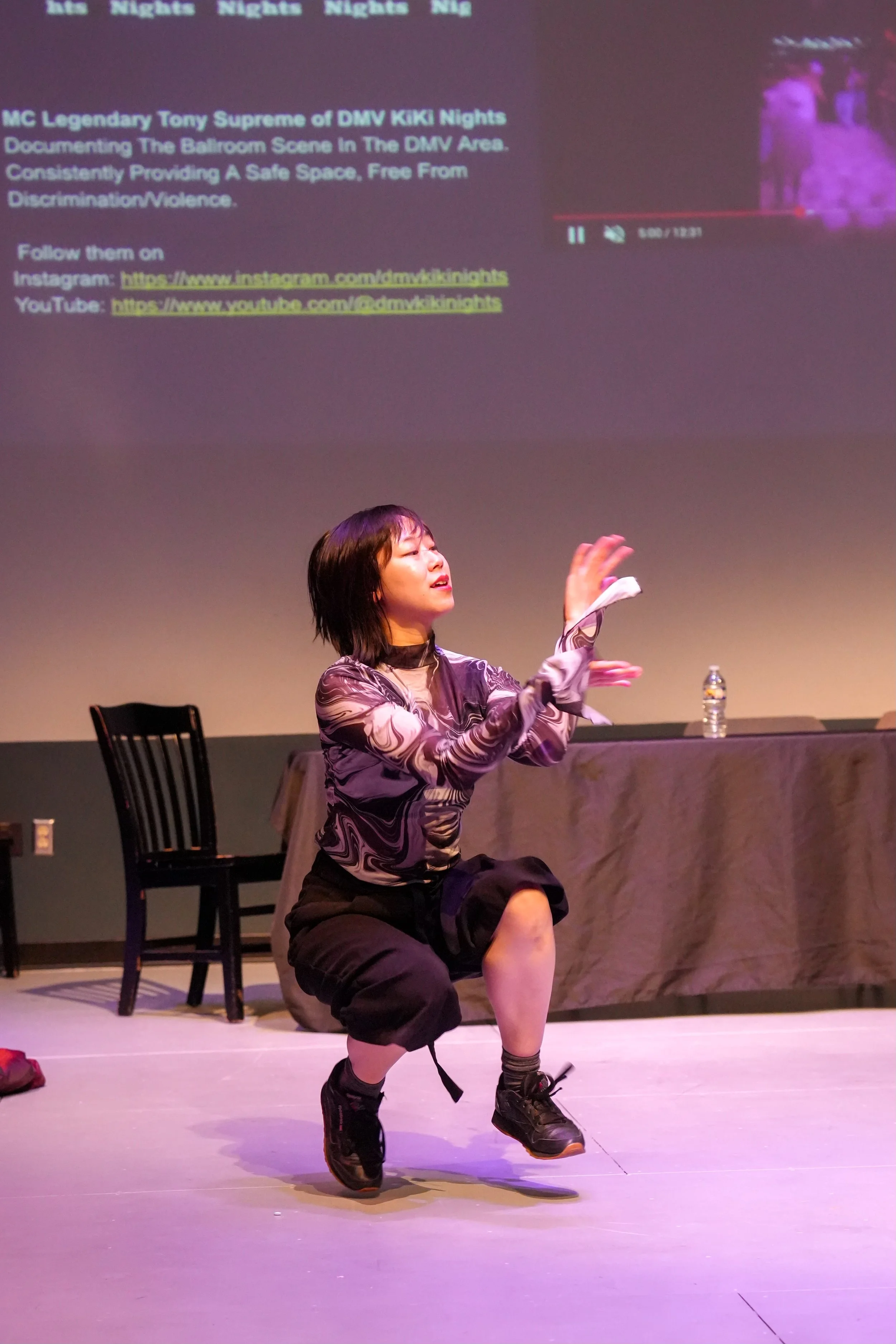 A woman performing a dance move on stage, kneeling with one arm raised and the other bent at her side, wearing a patterned top, shorts, and sneakers, with a large projection screen in the background displaying text and a video preview.