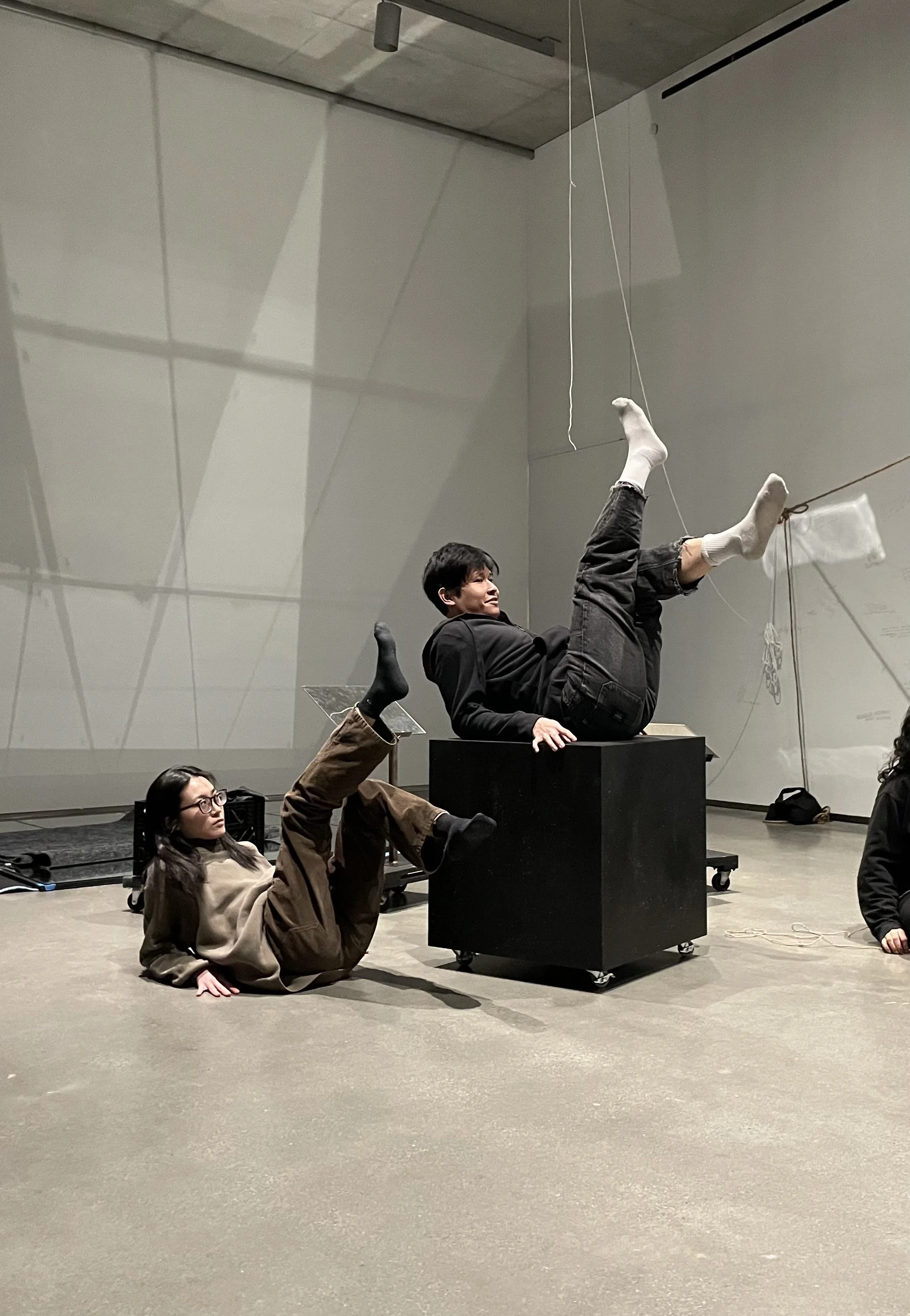 People engaging in an artistic or theatrical performance or activity in a modern, minimalistic indoor space. One person is lying on a box, with their legs raised and extended, while another person is lying on the floor nearby, supporting themselves with one arm and wearing glasses.