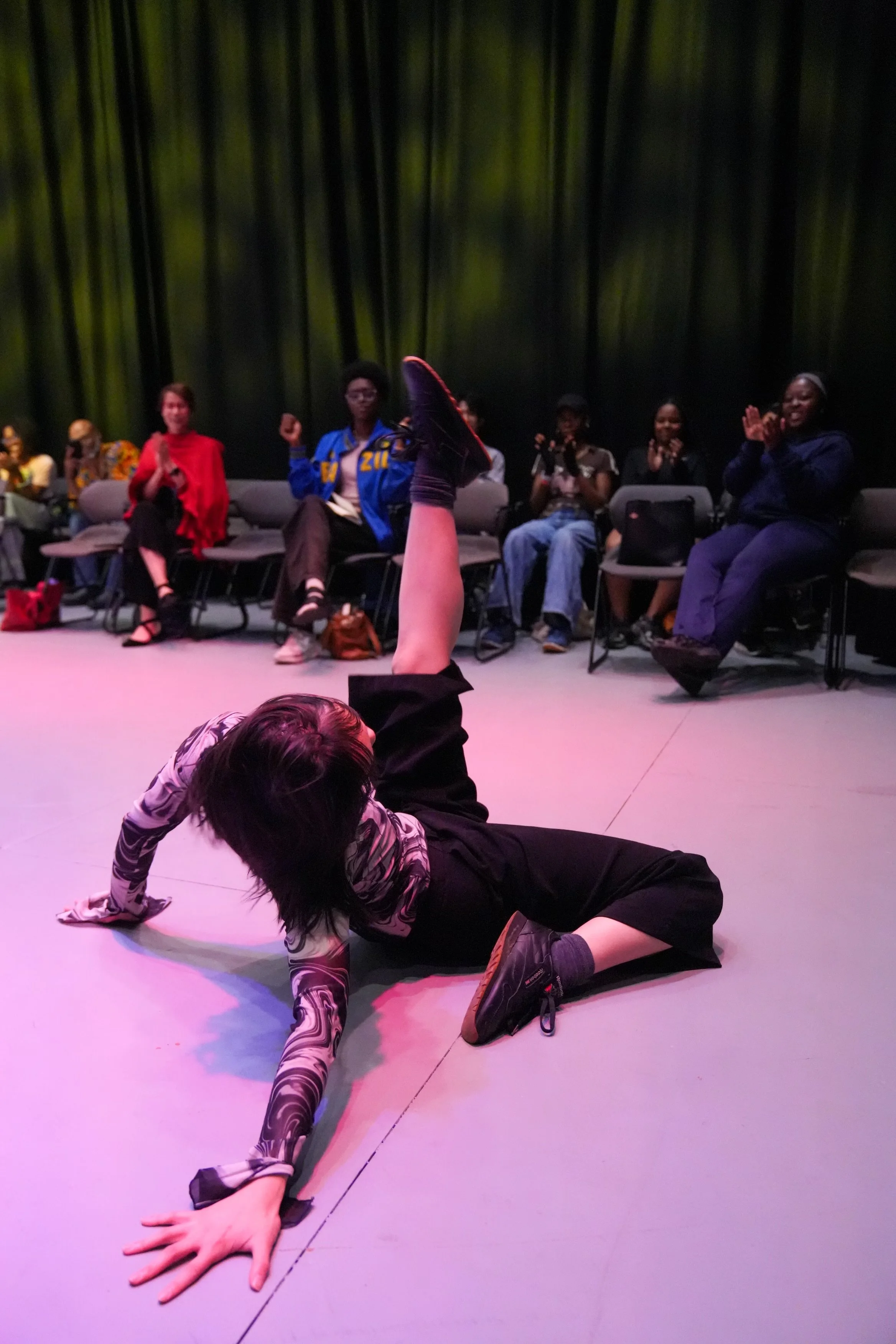 A person lying on the floor with one hand supporting their body and the other arm extended, while looking towards an audience seated on chairs. The audience, mostly women, are clapping and smiling, in a dark setting with a green curtain backdrop.