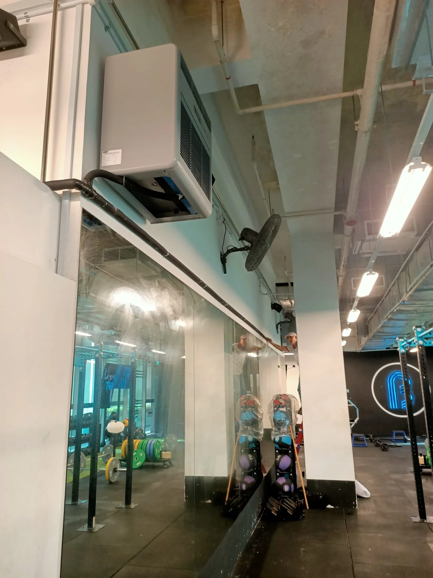 Inside a gym with equipment, kettlebells, and medicine balls visible through a glass wall. Two people are working on a ceiling dehumidifier installation, with tools and supplies nearby.