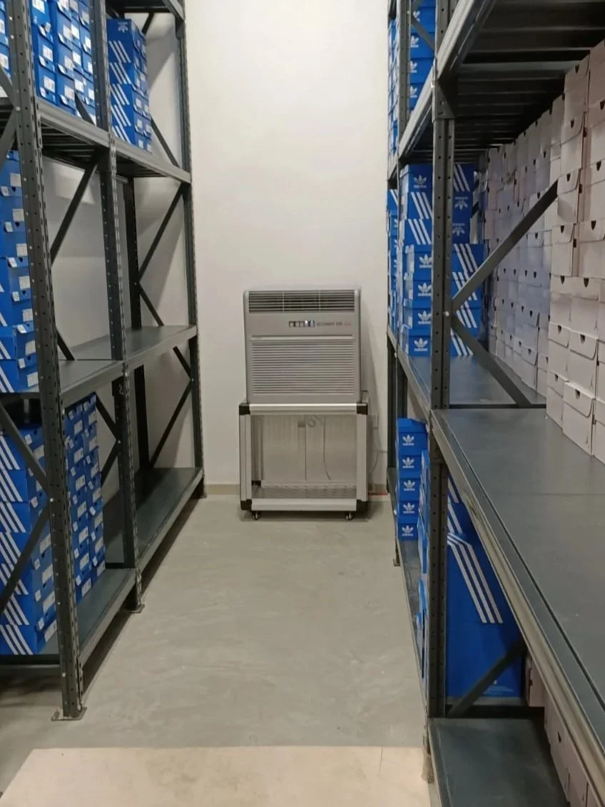 Empty storage room with metal shelving units containing blue and white boxes, dehumidifier unit, and a metal cart.