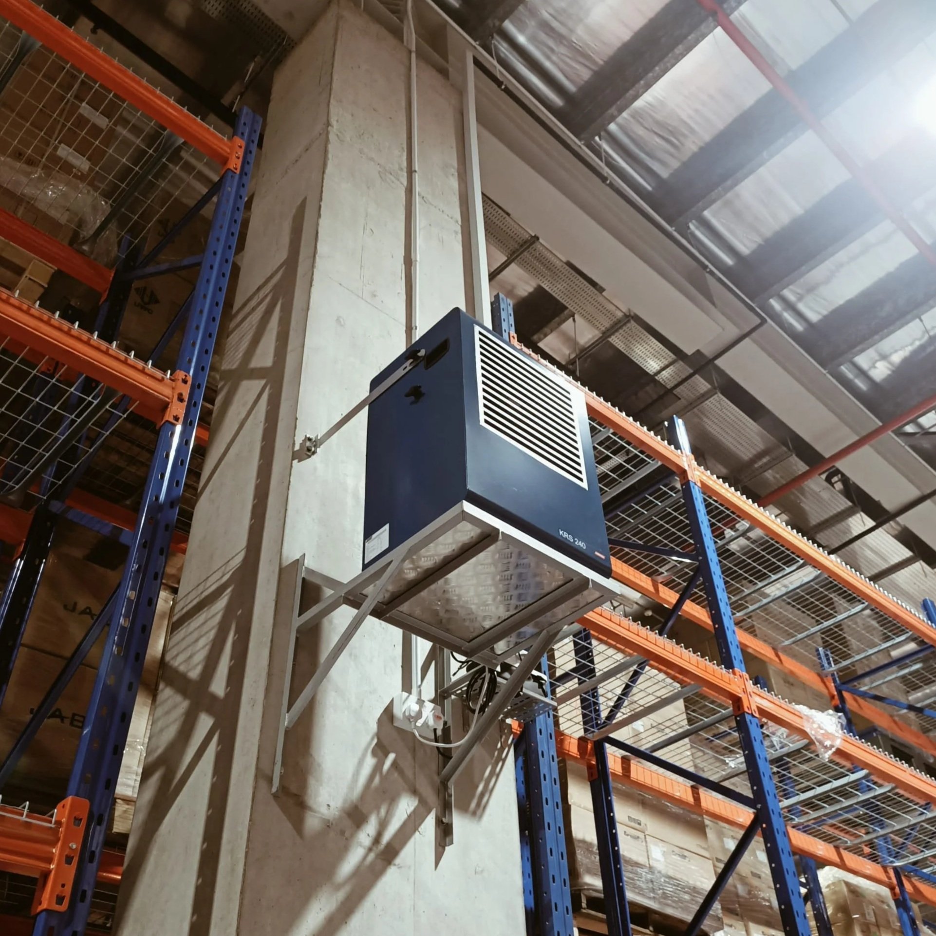 Industrial warehouse with blue and orange metal shelving, a blue dehumidifier attached to a concrete wall, and metal ceiling with skylights.