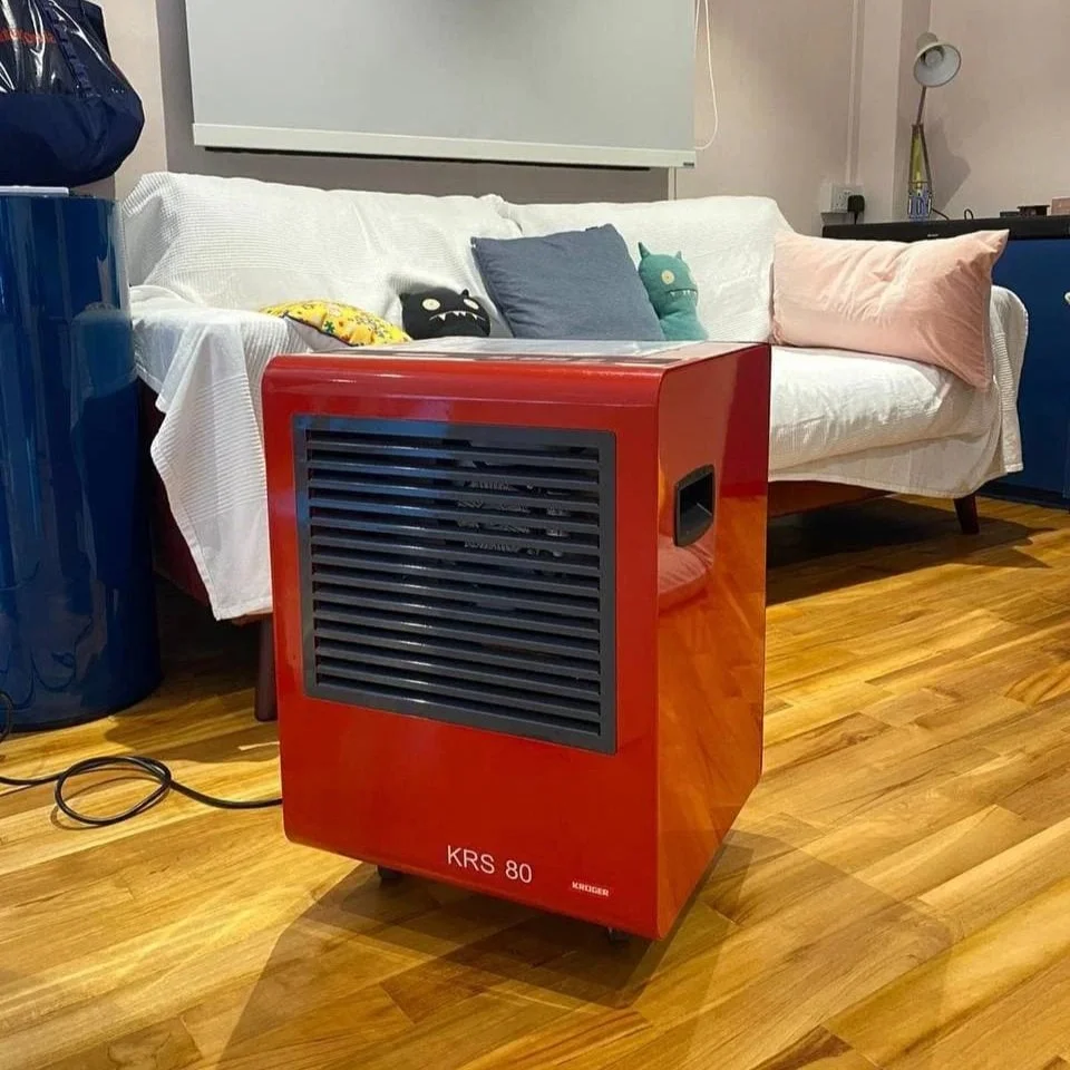 A red dehumidifier labeled 'KRS 80' on a wooden floor in a living room. Behind it is a white couch with multiple pillows and stuffed toys, along with a blue container and wall-mounted lights.
