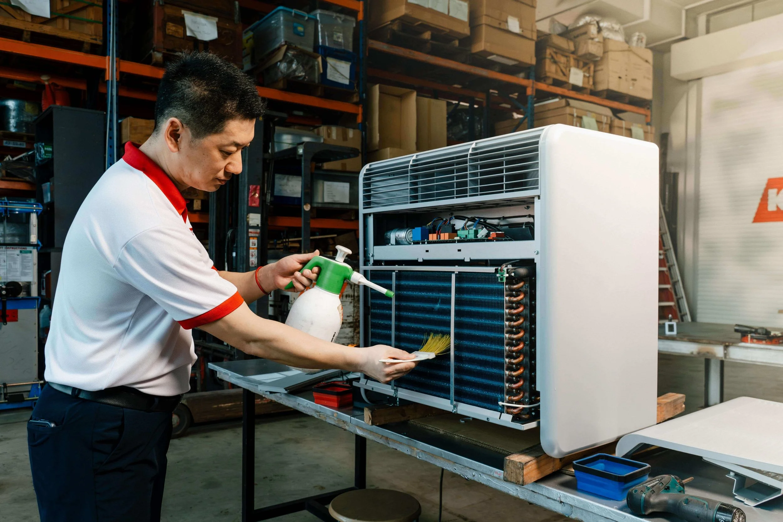 A technician cleaning the interior components of a dehumidifier unit with a spray bottle and a brush in a warehouse.