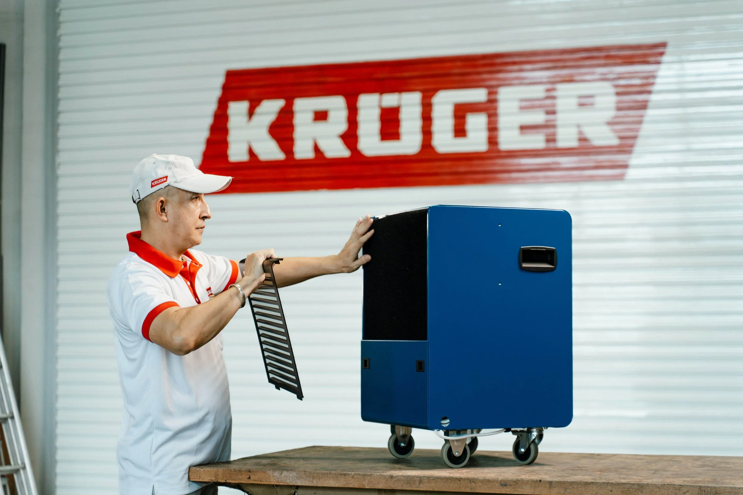 A man in a Kruger uniform and cap places a vent cover into a dehumidifier unit on a table, with a Kruger sign in the background.