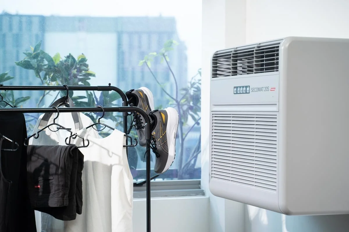 Clothes hanging on a drying rack next to a dehumidifier unit by a window with plants outside.