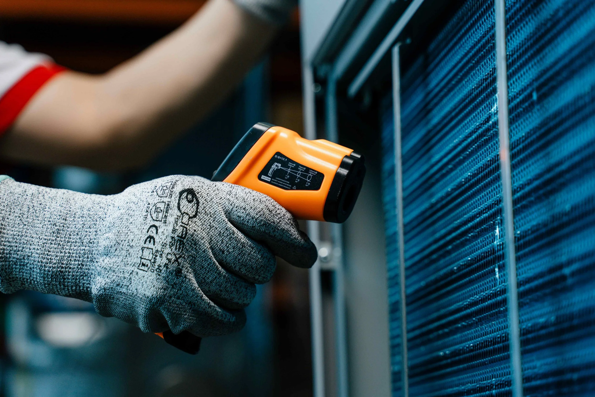 A person wearing a grey glove with safety markings is using a handheld infrared thermometer against a dehumidifier.