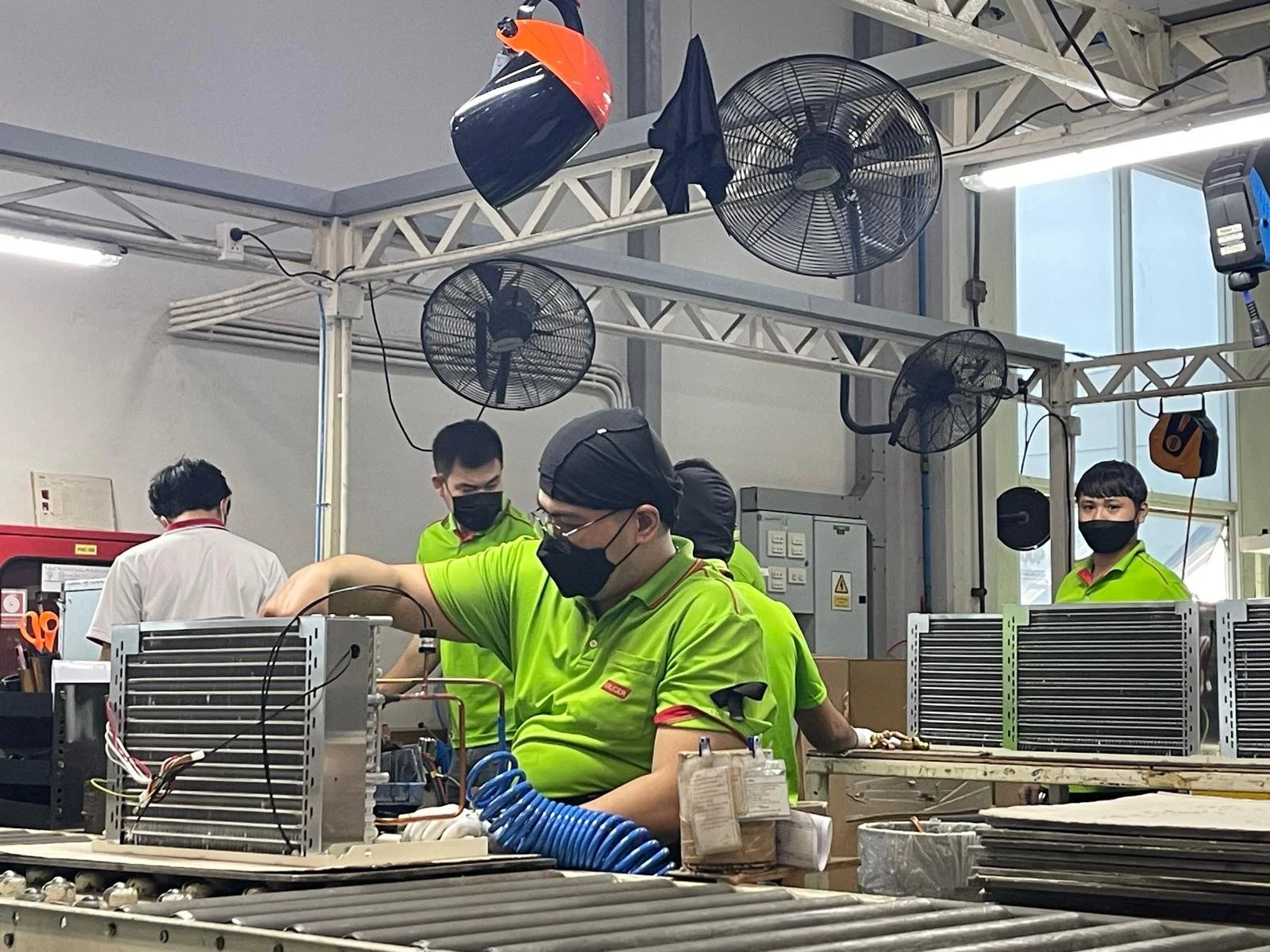 Workers in a factory assembling or repairing electronic equipment, wearing bright green uniforms and black masks, surrounded by electronic components and tools, with industrial fans visible on the ceiling.