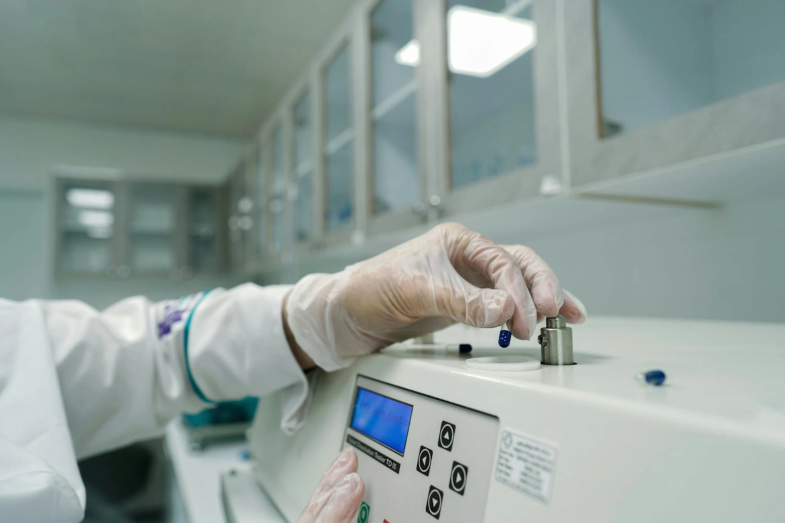 A lab technician wearing gloves handling blue capsules in a laboratory setting, adjusting a machine with a digital display.