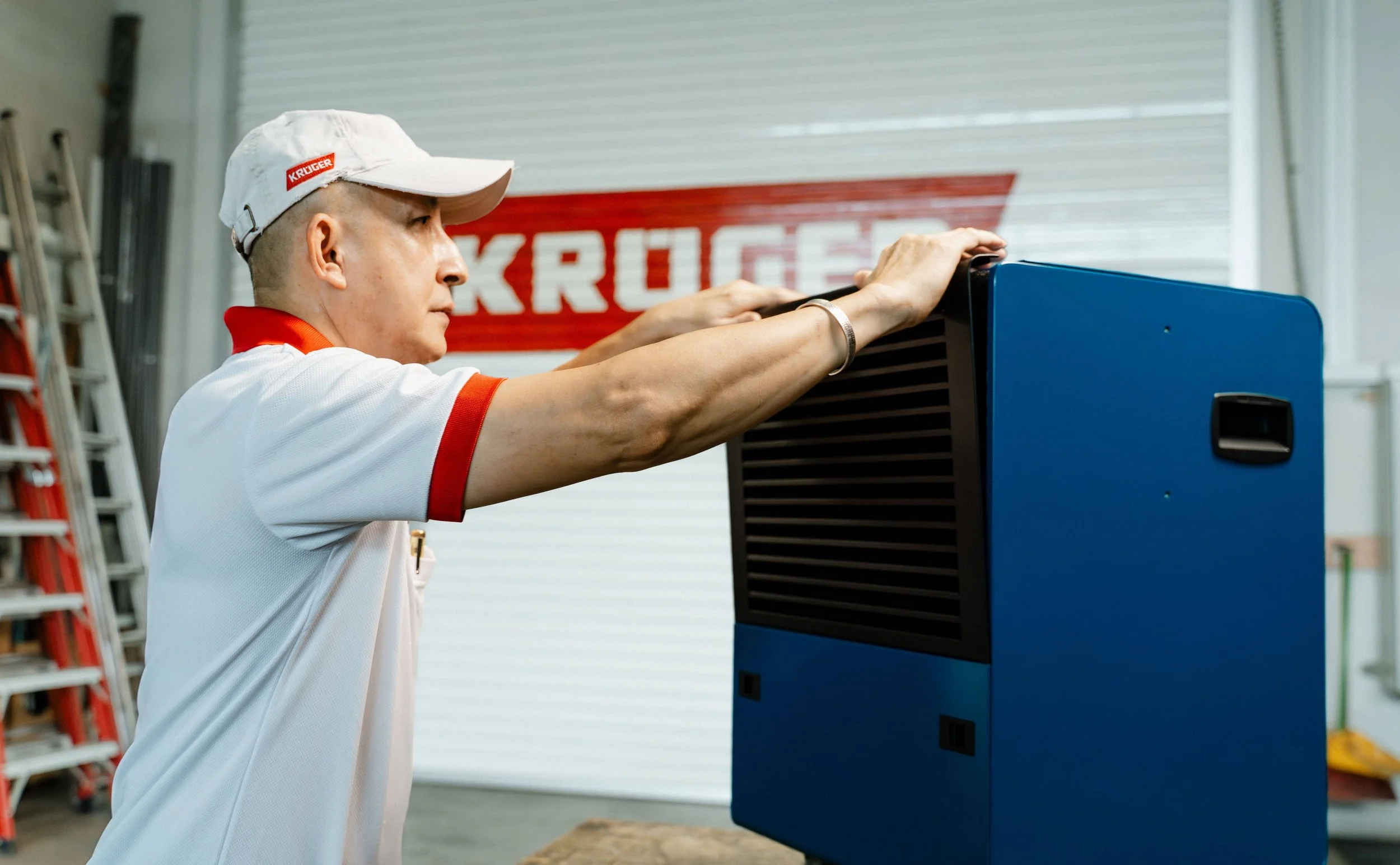 A man in a Kruger uniform and white cap is working on a blue Kruger Dehumidifier unit inside a Kruger warehouse. The background features a Kruger sign and some ladders.
