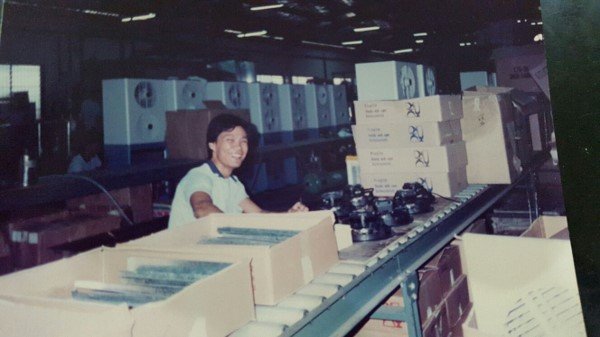 A young boy sitting at a counter with electronic equipment and boxes, in an indoor warehouse, with dehumidifiers visible in the background.
