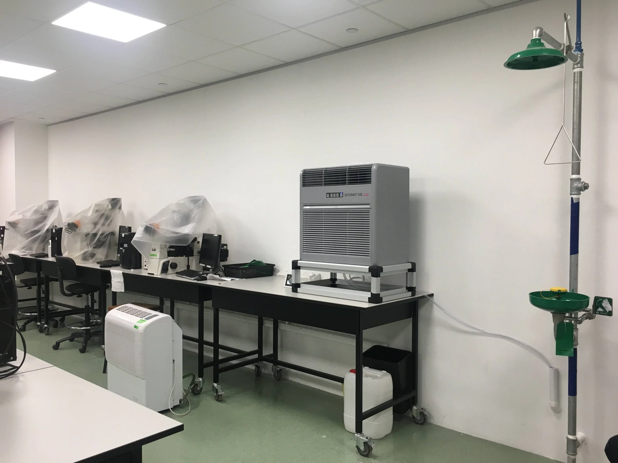 Laboratory or cleanroom with scientific equipment, computer stations, and a large machine on a cart. There is a green and white medical or scientific device on wheels, and a makeshift sink with a green basin and faucet attached to a pole. The room has white walls and a ceiling with fluorescent lights.