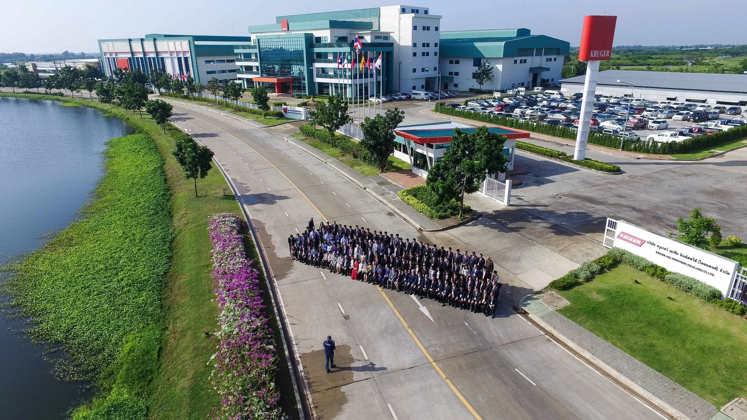 Large group of people standing on a wide road in front of a large corporate building with a parking lot and a tower with the brand name Kruger, near a lake with green water plants.