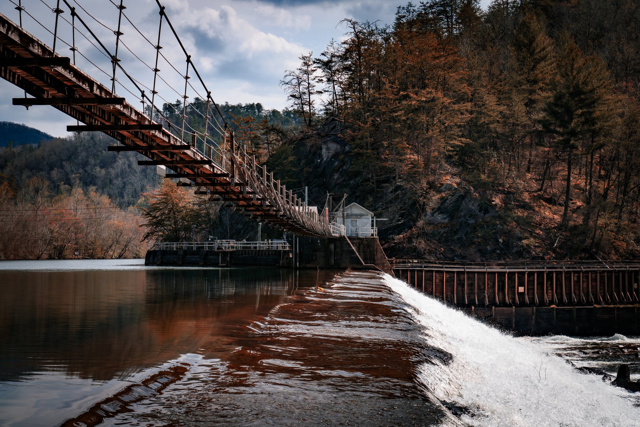 A suspension bridge over a body of water with trees and rocky hills in the background.
