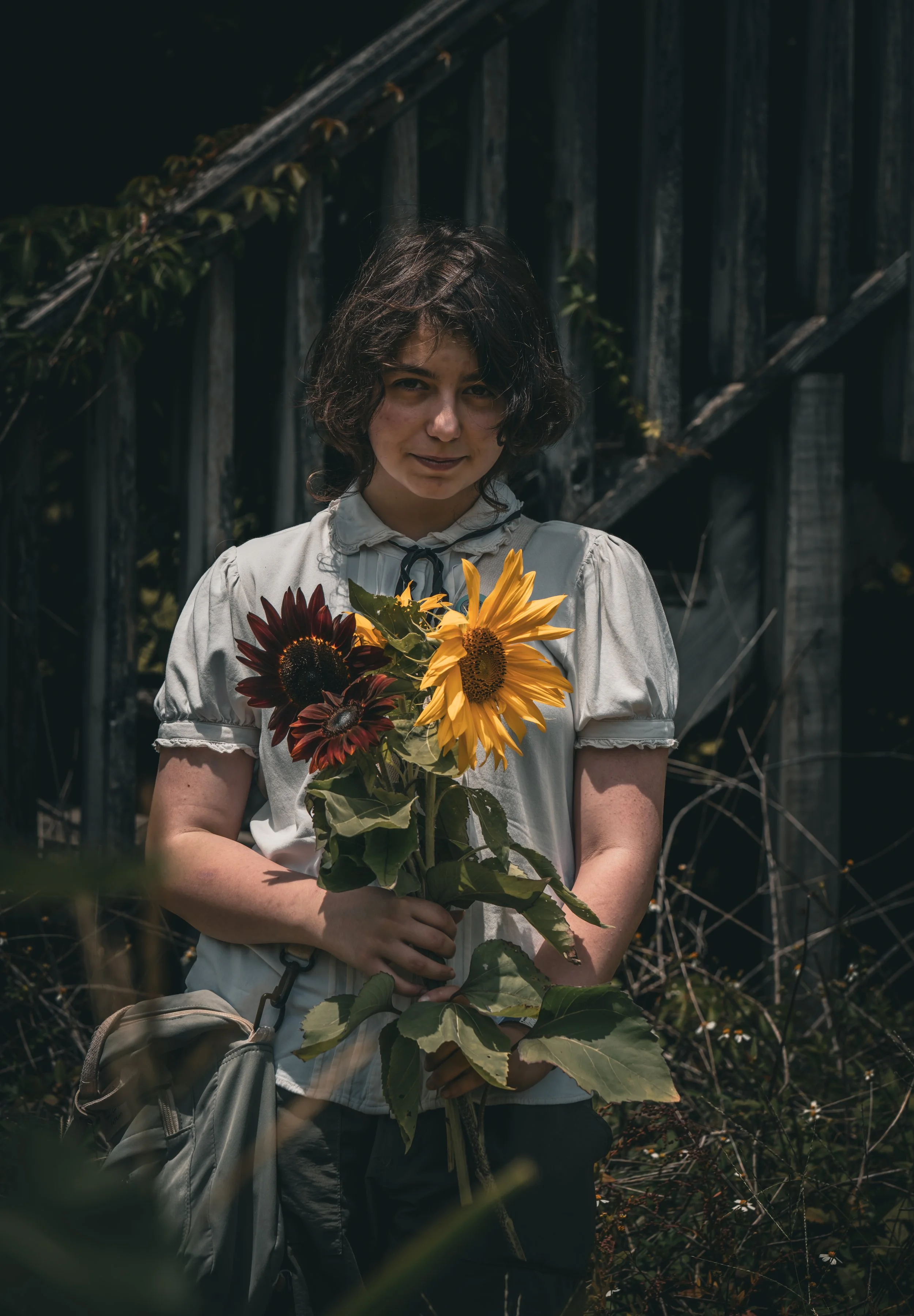 A woman with short, curly brown hair standing outdoors in front of a weathered wooden fence, holding a bouquet of large, vibrant sunflowers and dark red flowers, wearing a light-colored blouse.