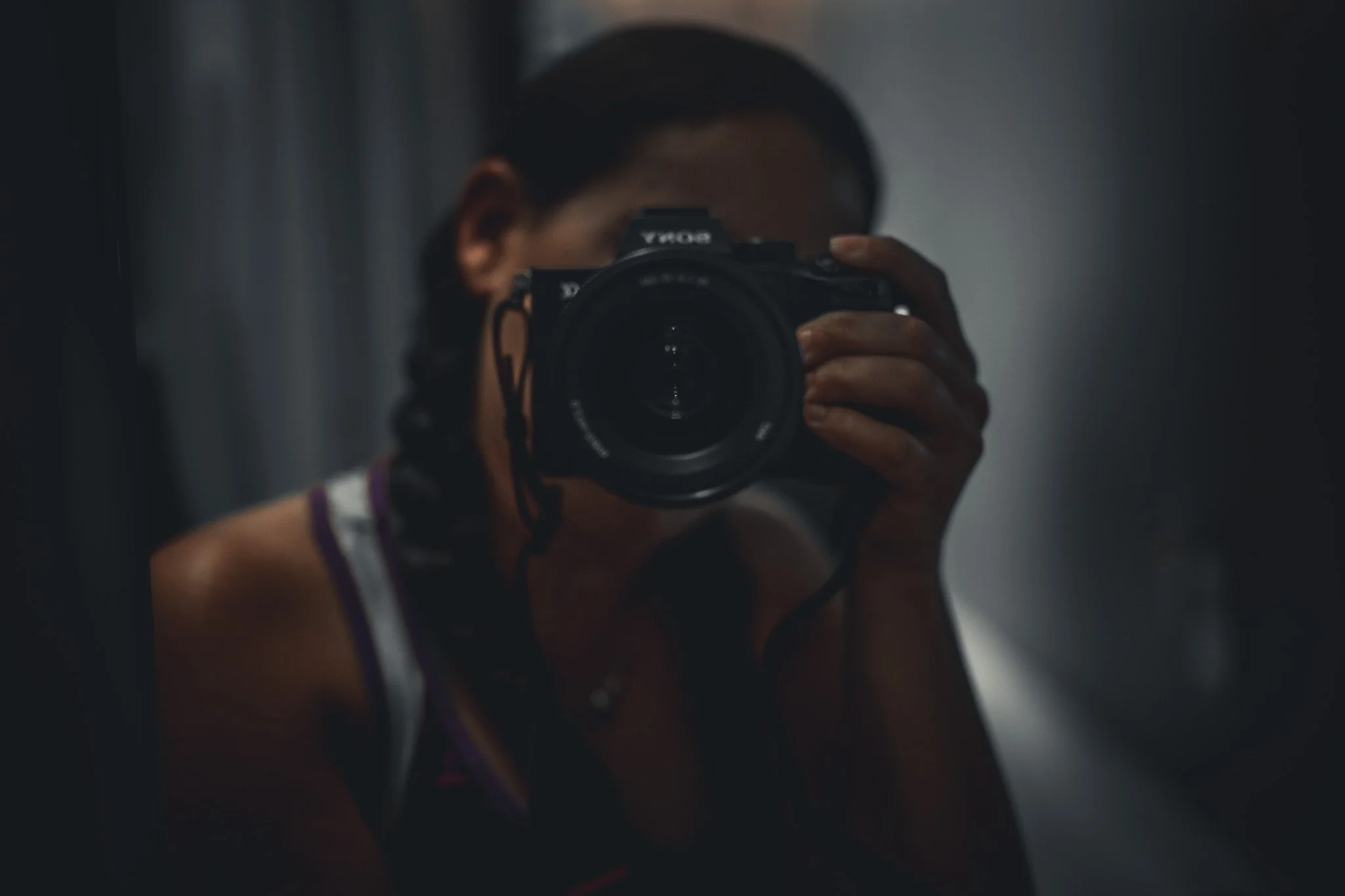 Person taking a selfie in a mirror with a camera, dark lighting, woman with braided hair wearing sports top.