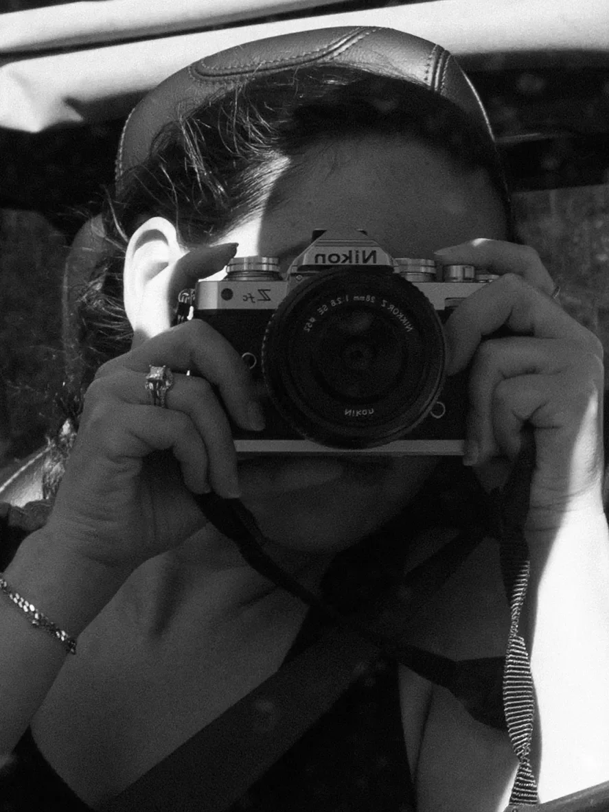 A person taking a selfie with a Nikon camera, wearing a hat and jewelry, reflected in a mirror.