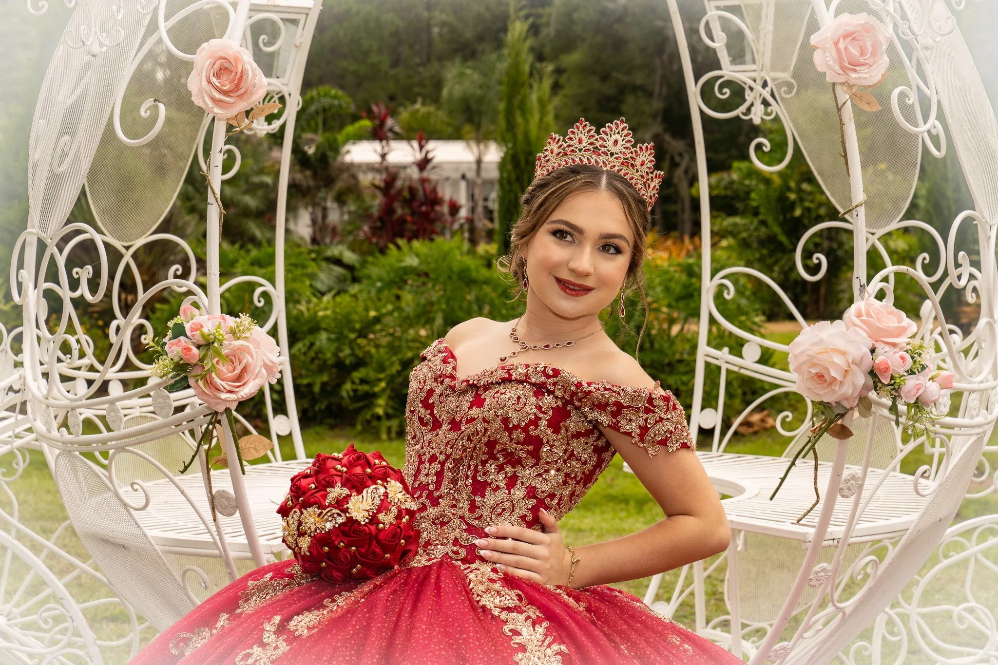 Quinceañera photo. Young woman in a red and gold embroidered gown with a matching tiara sitting on a white iron garden swing decorated with pink roses, outdoors in a lush garden.