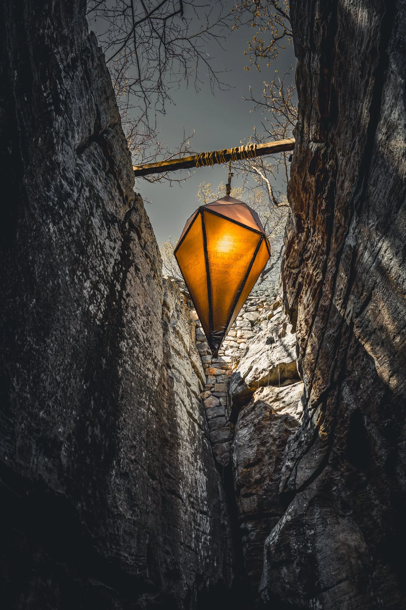 A lit orange lantern hanging between rocky canyon walls, with a partly cloudy sky and barren tree branches overhead.