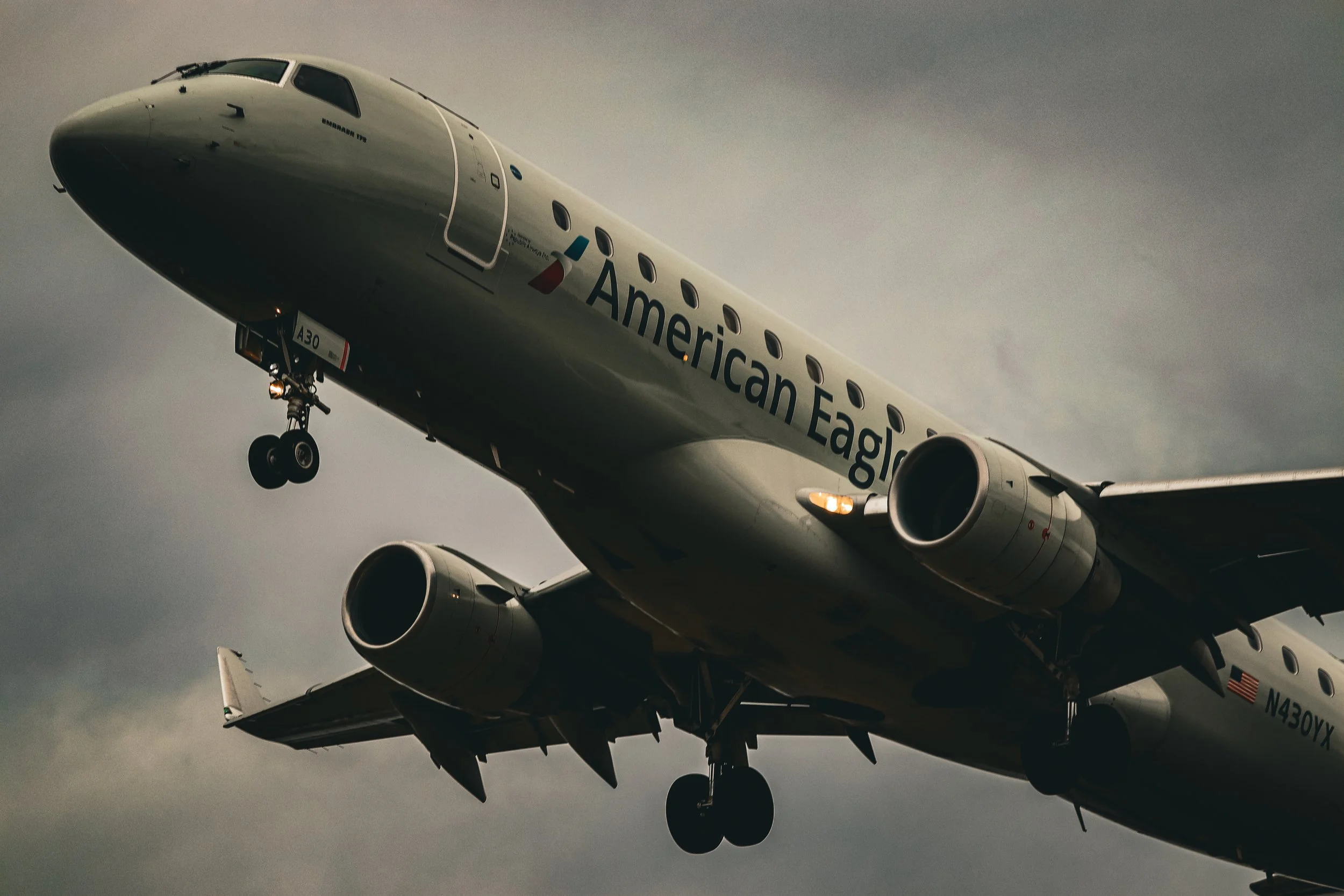 An American Eagle airplane in flight against a cloudy sky, with landing gear deployed and engines visible underneath the wings.