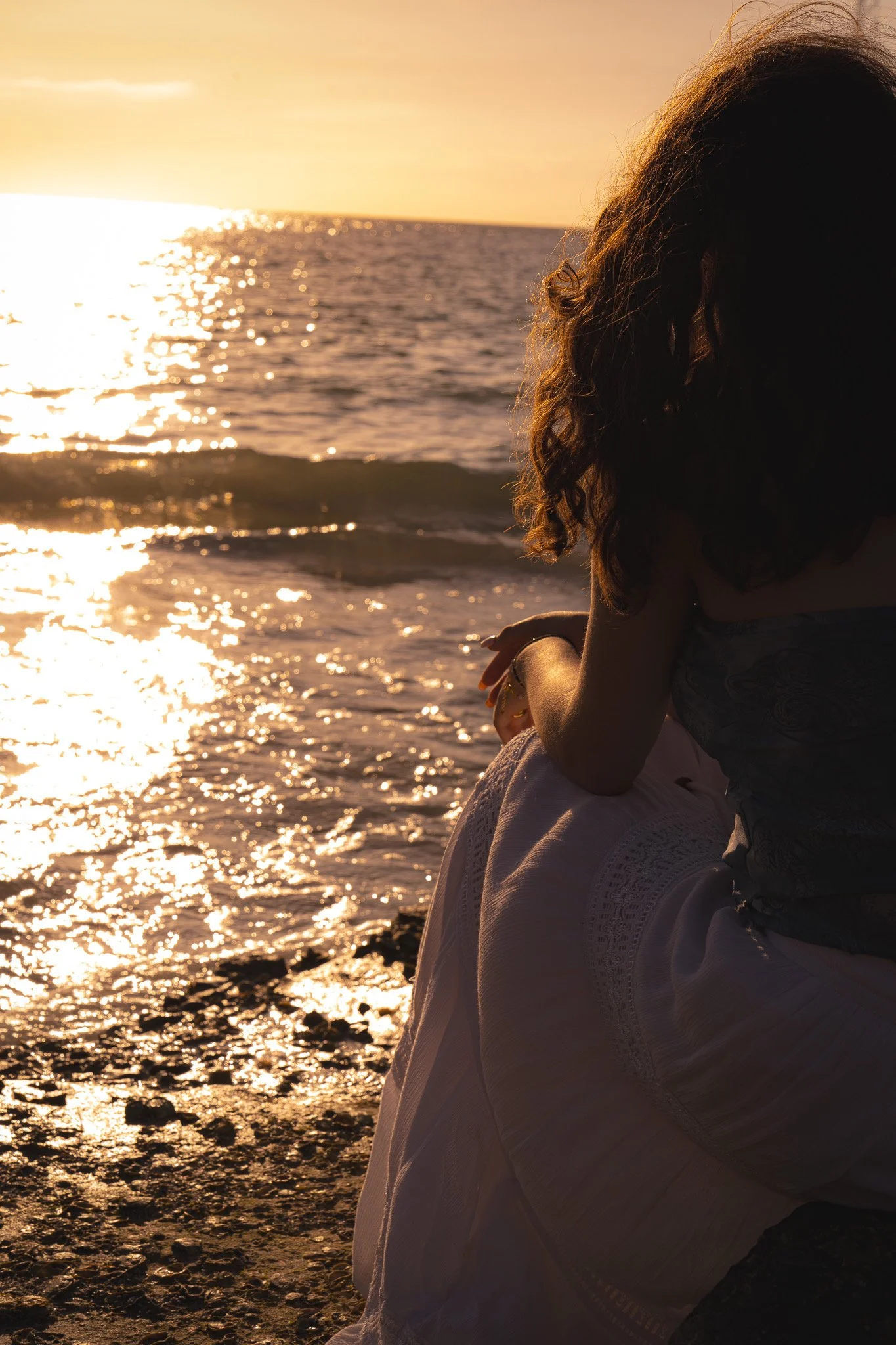 A woman sitting on a beach at sunset, looking out towards the ocean, with sunlight reflecting on the water.