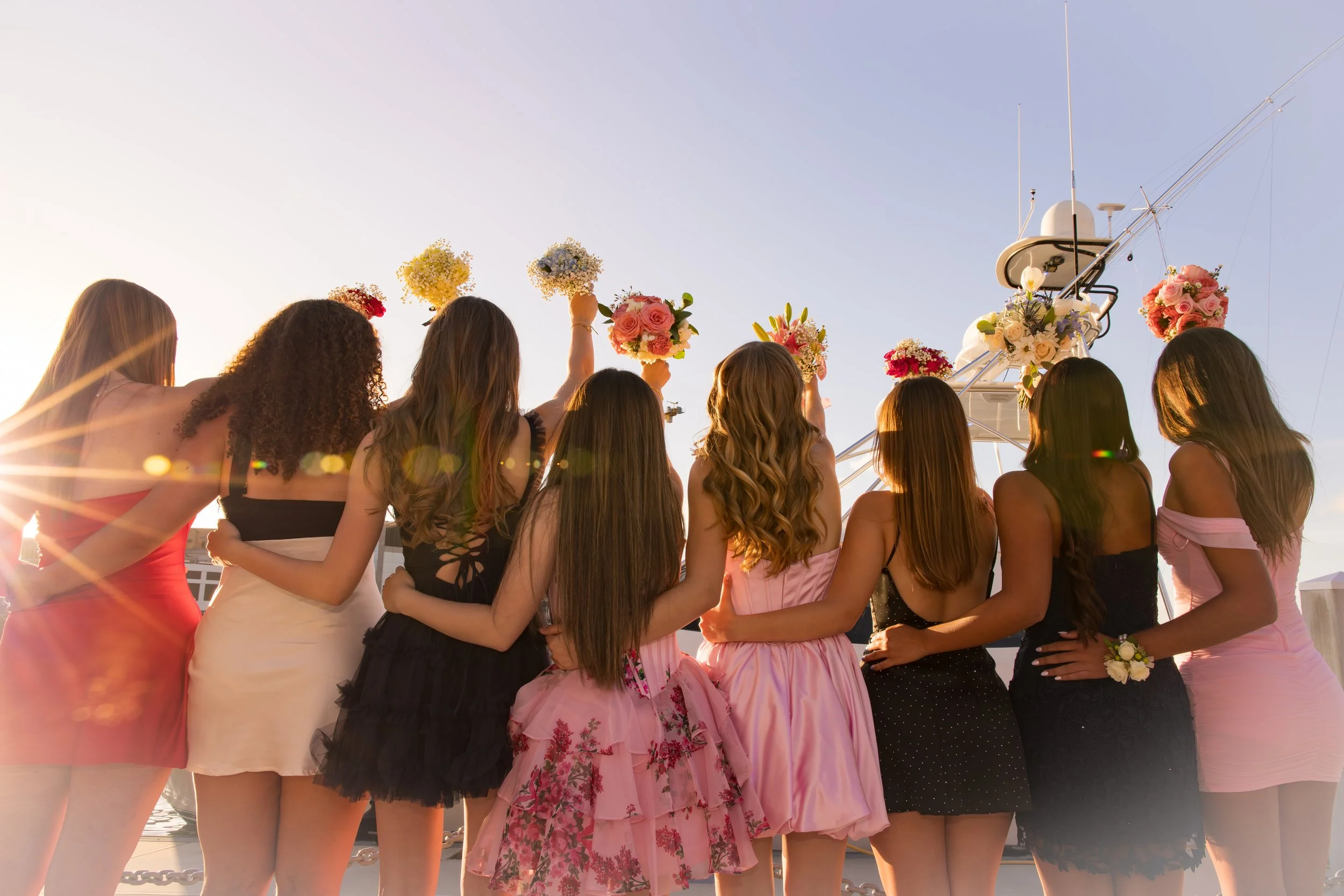 Group of young women dressed in colorful dresses, holding bouquets, standing on a dock near a yacht at sunset.
