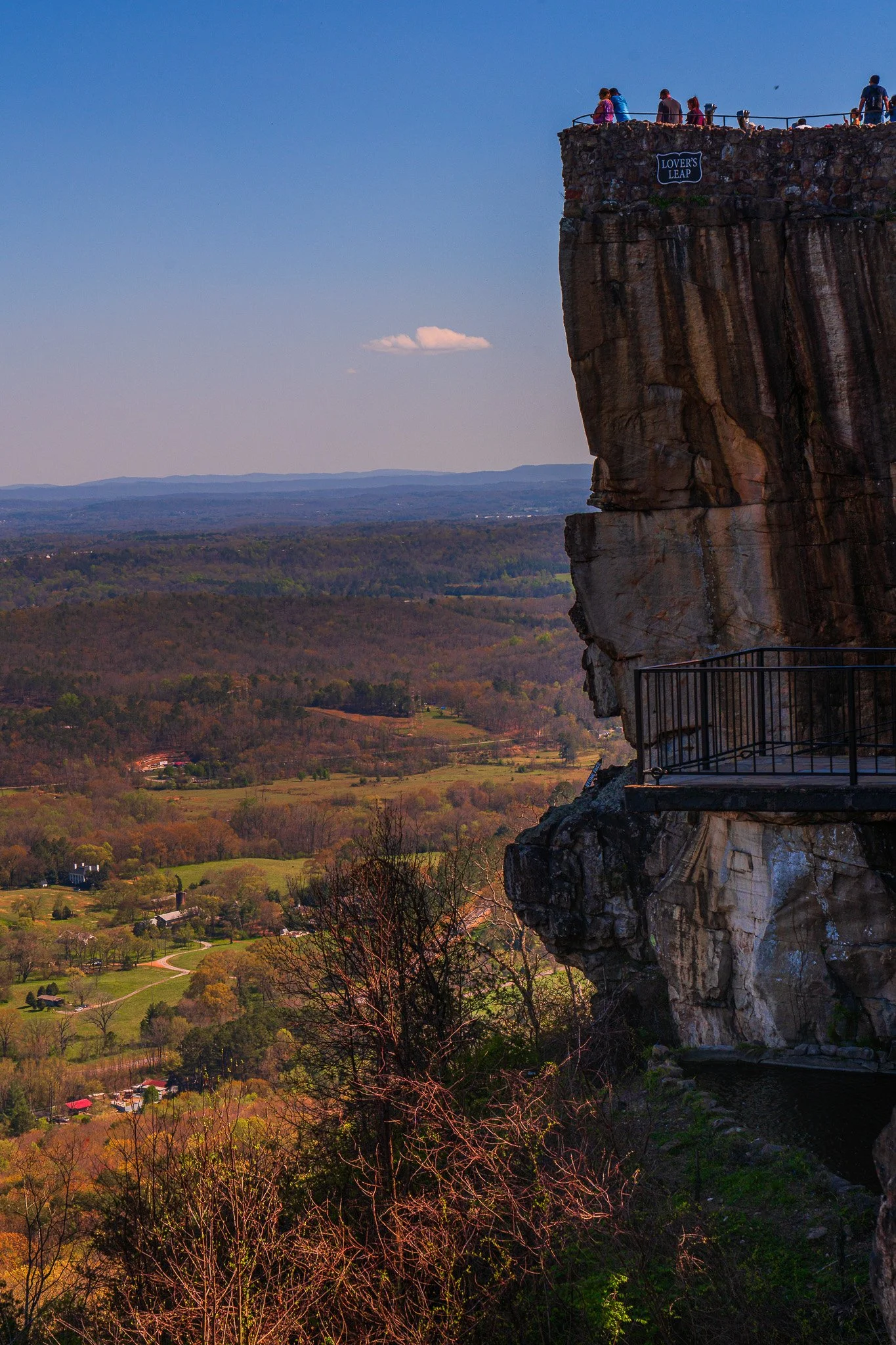 People standing on an observation deck on a rocky cliff called Lover's Leap, overlooking a broad landscape with trees, fields, and distant mountains on a clear day.