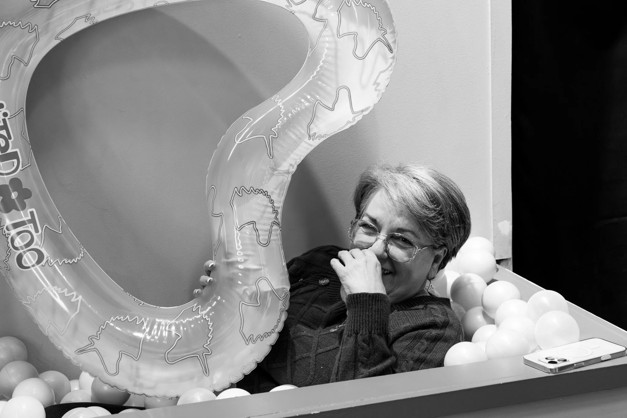 A woman with glasses smiling and laughing while sitting in a ball pit with a large inflatable heart and a smartphone nearby.