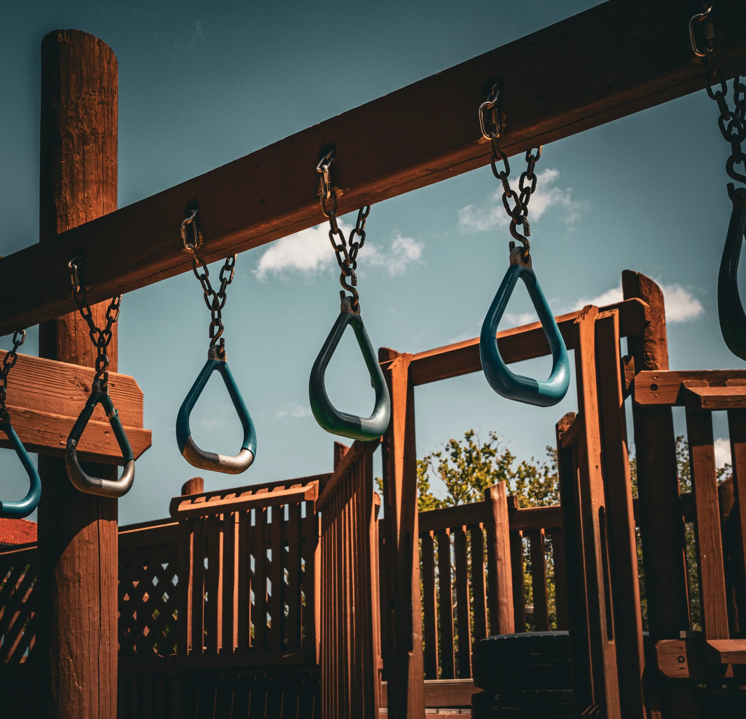 Close-up of a wooden playground structure with blue and black swings hanging from chains, under a partly cloudy sky.