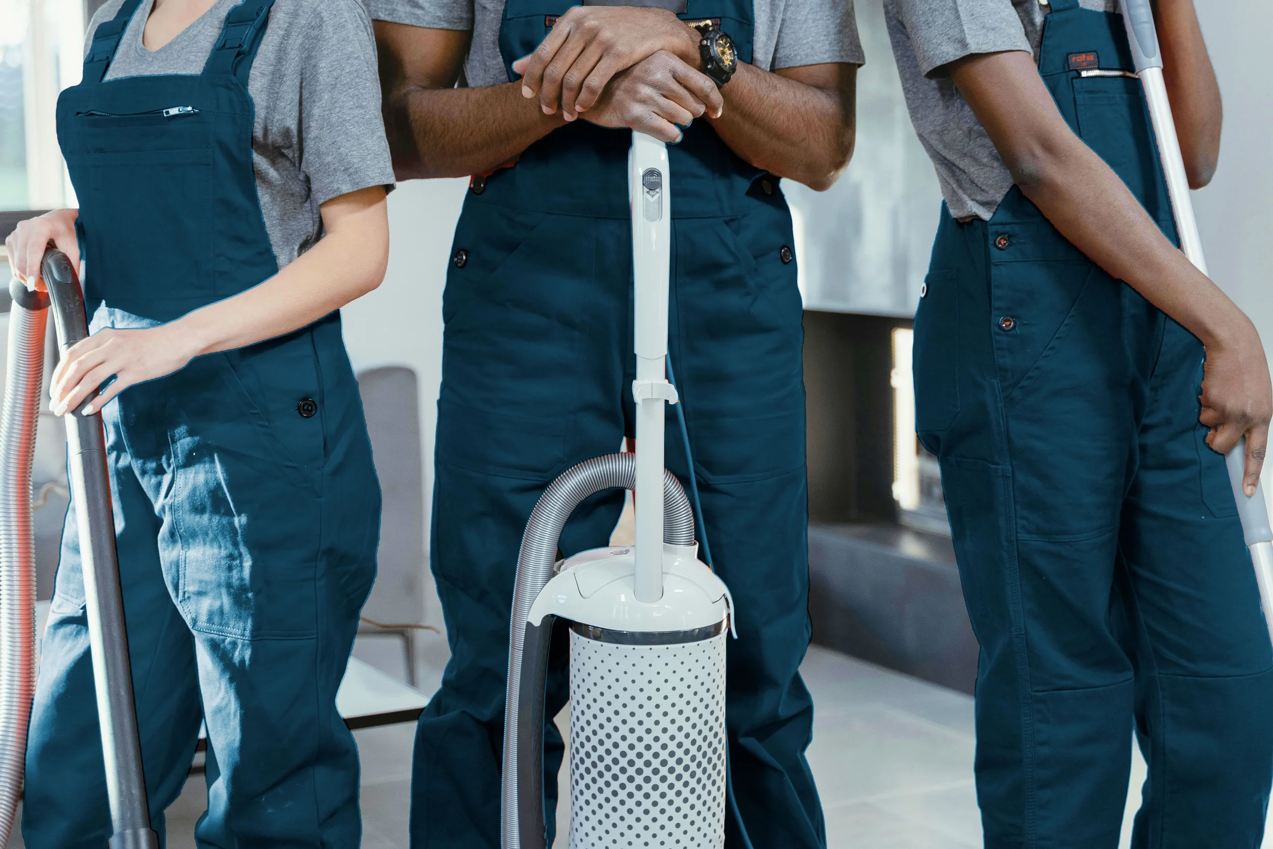 Three people in gray shirts and blue work pants holding vacuum cleaners, standing side by side in a room.