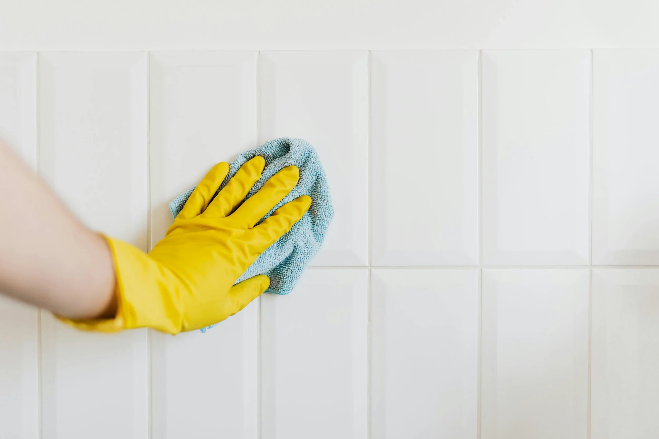 Person wearing yellow gloves cleaning white tiled wall with a cloth.