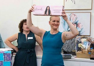 A woman in a blue sleeveless top holding up a pink box, possibly a product, in a store or boutique. Another woman stands nearby, observing. The background features framed artwork and shopping bags.